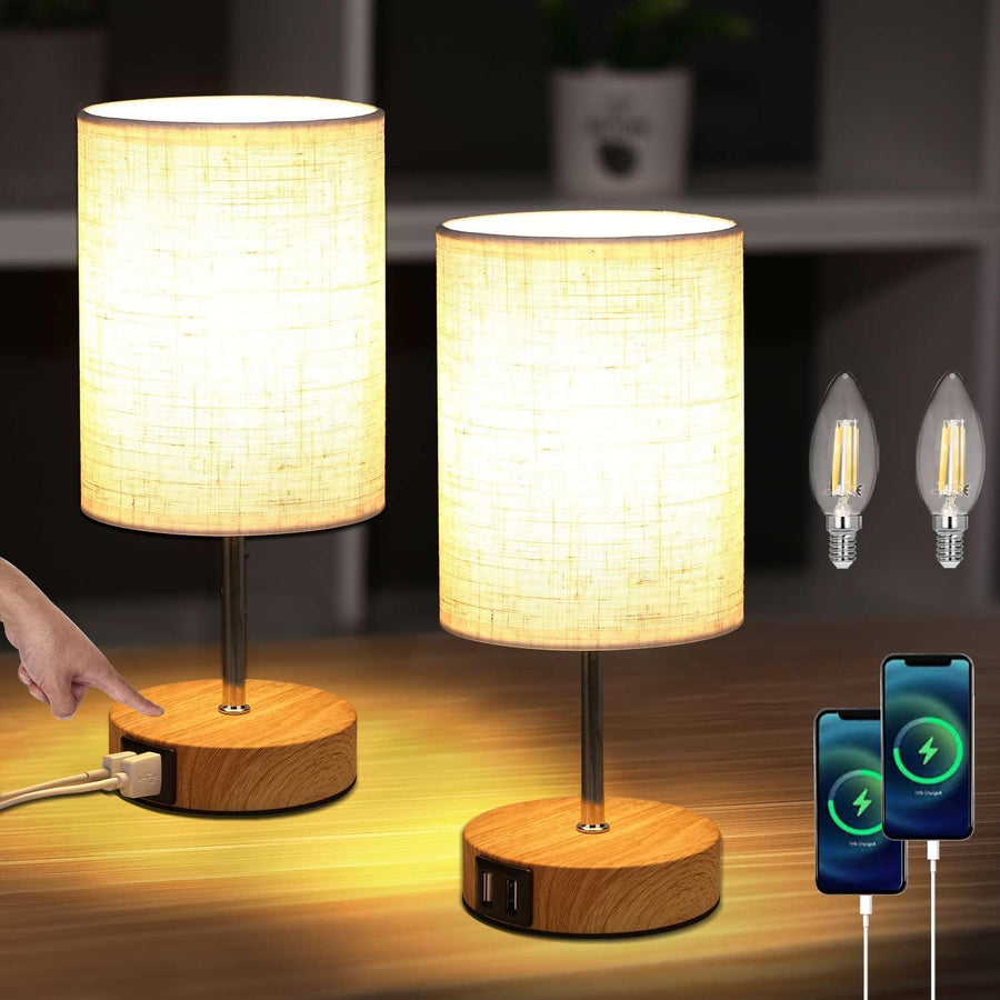 Two table lamps with wooden bases and fabric shades, one being turned on, with two light bulbs and a smartphone charging cable in the background.
