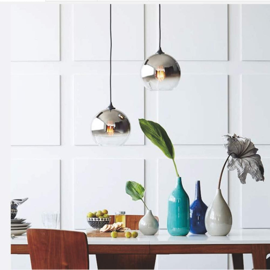 Modern pendant lights with silver glass shades and dark cord, placed over a white kitchen cabinet, with decorative vases and a plant on the table below.