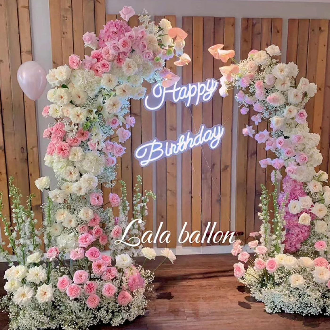Floral arch with 'Happy Birthday' neon sign and 'Lala balloon' text against a wooden background.