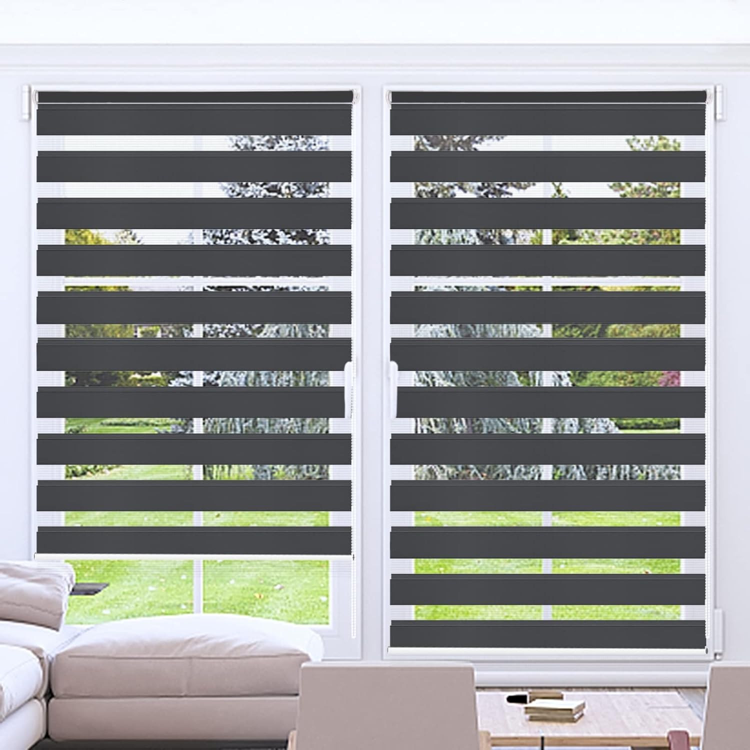 A pair of dark grey zebra roller blinds with horizontal stripes, installed on a window with a view of a grassy area outside.