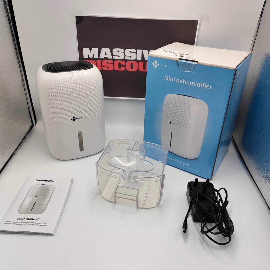 A white dehumidifier with its packaging box, a clear water container, and a power cord are displayed against a backdrop featuring 'MASSIVE SAVINGS' text. The product appears to be used with some signs of wear.