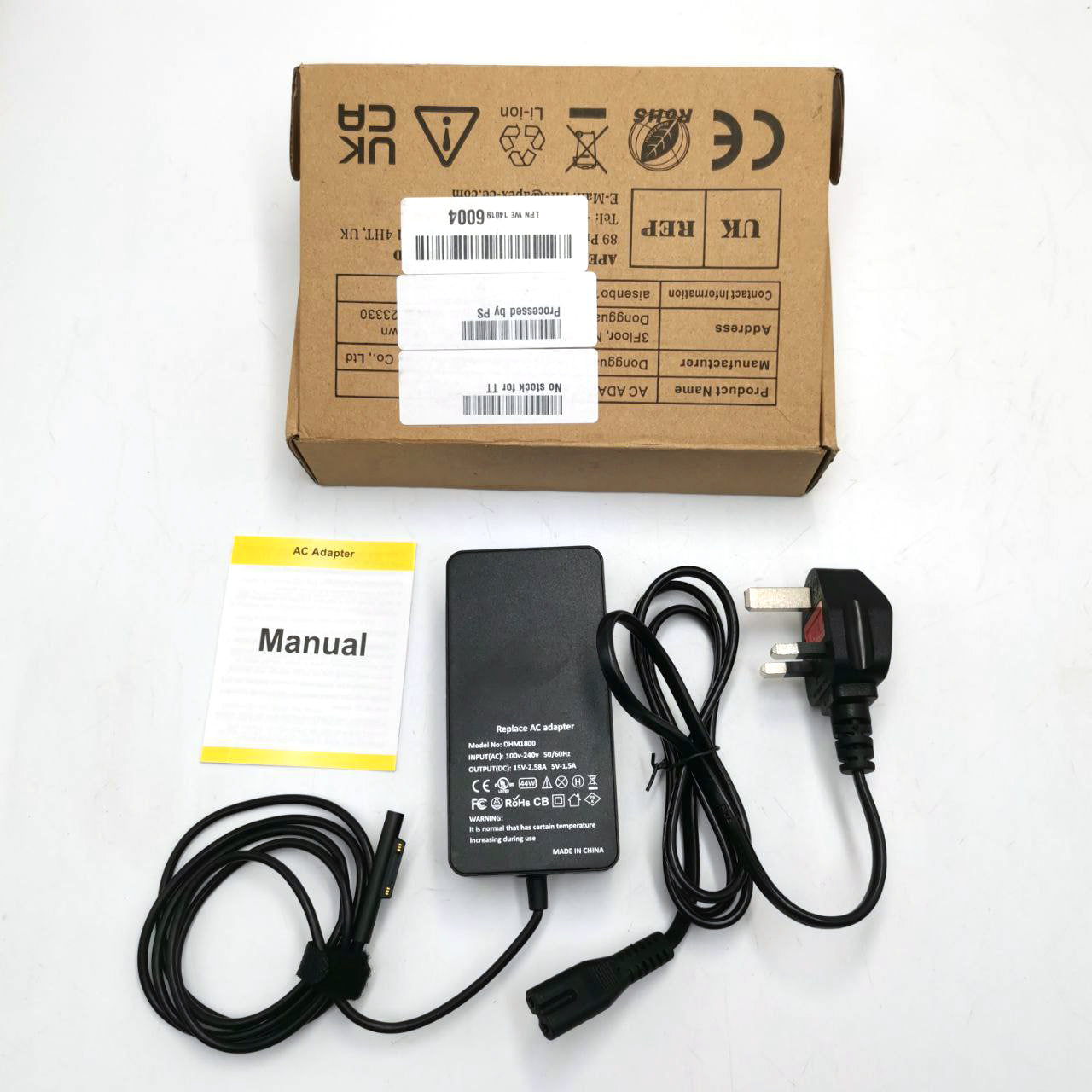 A black Microsoft Surface charger with a power cord, displayed next to its packaging box which has a label indicating 'Manual' and other details.