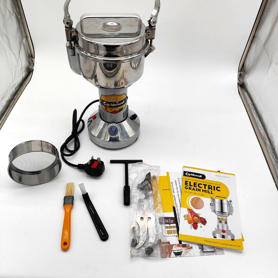 A silver electric grain mill grinder with accessories including a cleaning brush, a safety manual, and a pack of replacement parts, displayed on a white background.