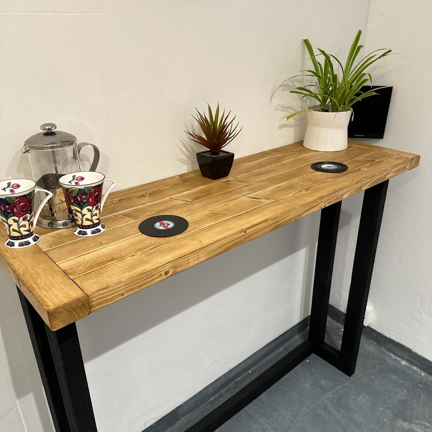 Wooden table with black metal legs, mugs, coasters, and plants on a white wall background.