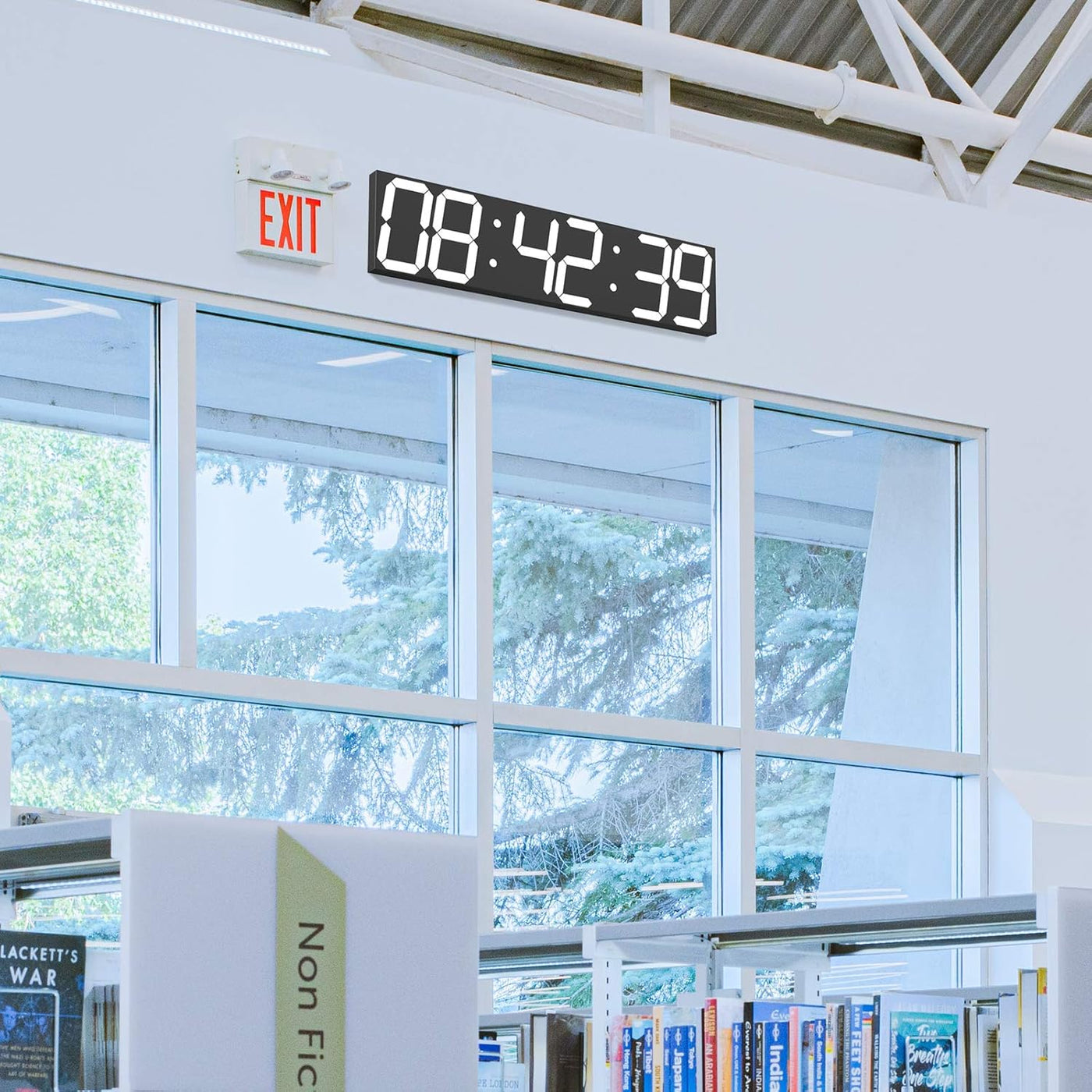 Digital clock displaying time in a library setting with books and large windows.