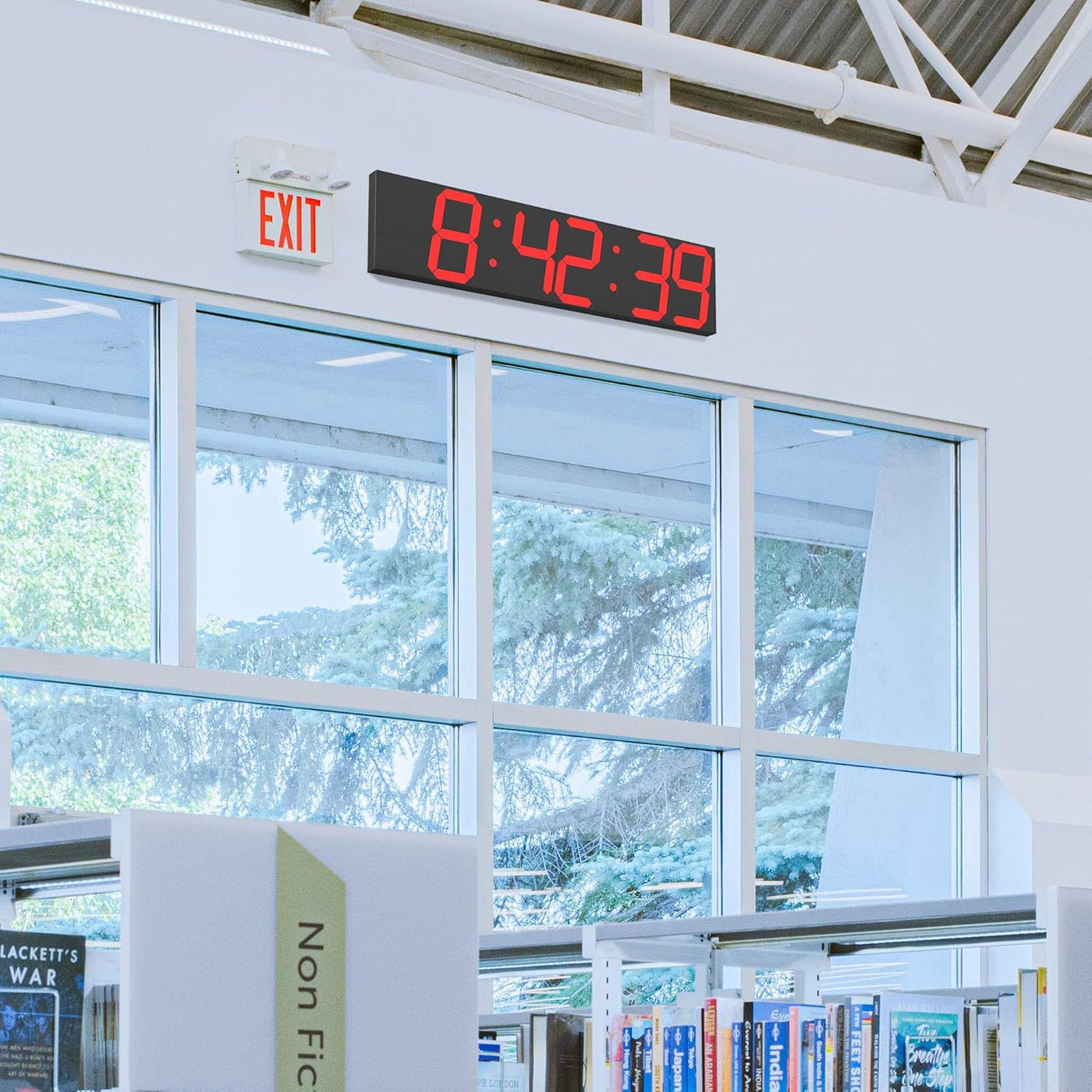 Digital clock displaying time in a library setting with books and large windows.