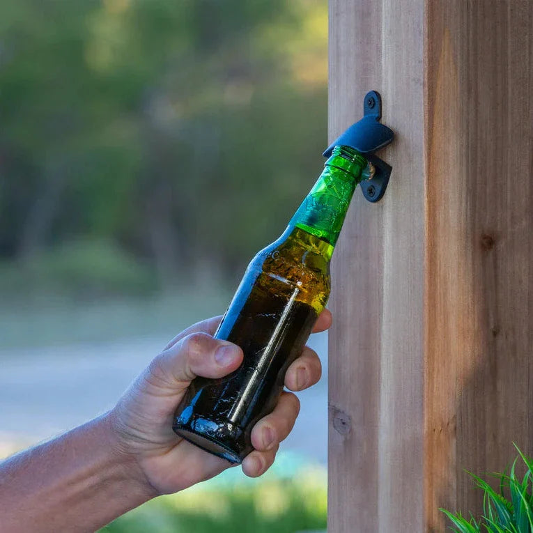 Hand holding a green bottle with a bottle opener attached to a wooden post outdoors.
