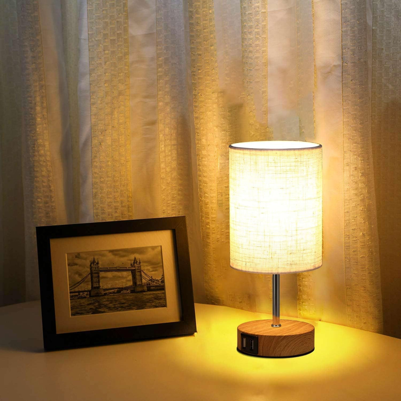 Table lamp with wooden base and fabric shade next to a framed picture on a surface with curtains in the background.