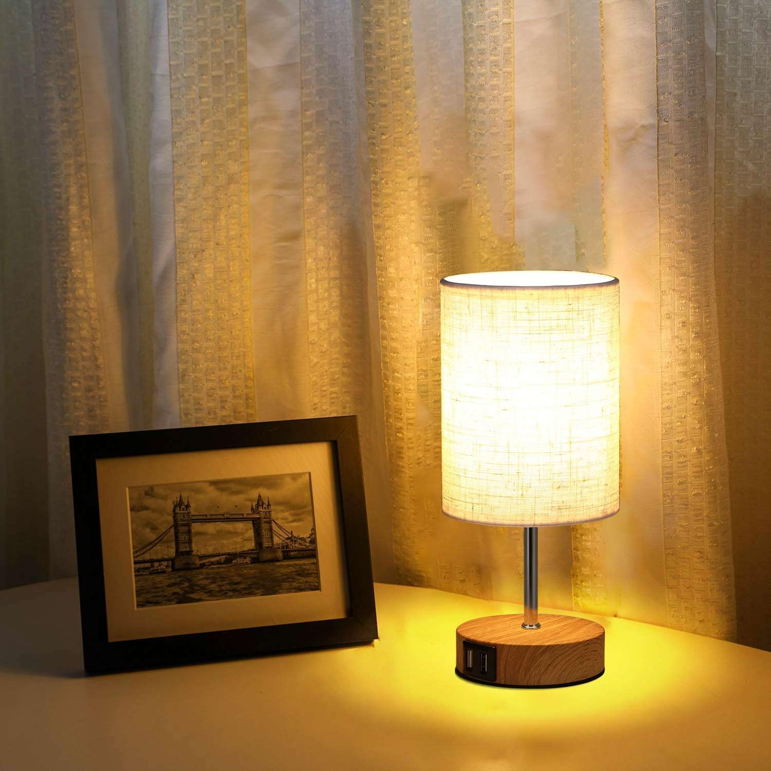Table lamp with wooden base and fabric shade next to a framed picture on a surface with curtains in the background.