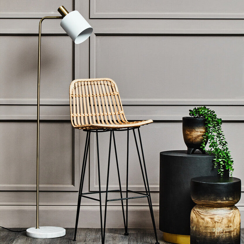 Wicker bar stool with black metal legs next to a floor lamp and small plant in a room with gray paneled walls.