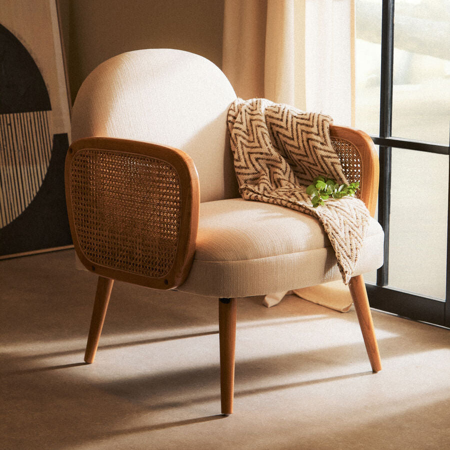 Beige armchair with wicker backrest and wooden legs, draped with a patterned blanket, in a softly lit room.