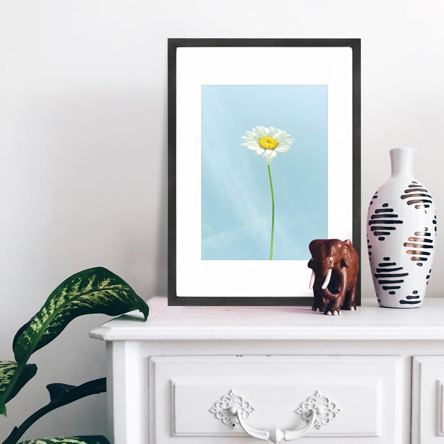 Framed artwork of a daisy on a white dresser with decorative items