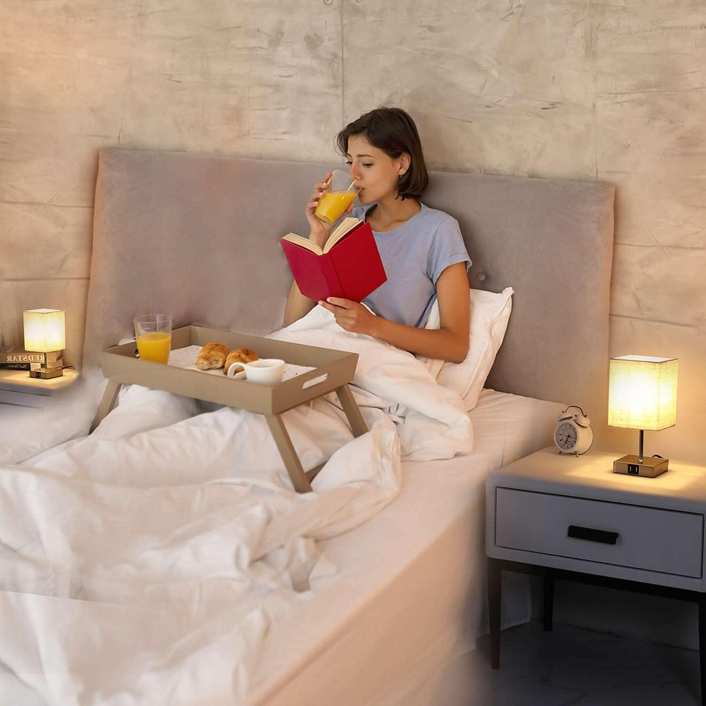 Woman reading a book in bed with a tray of breakfast and drinks.