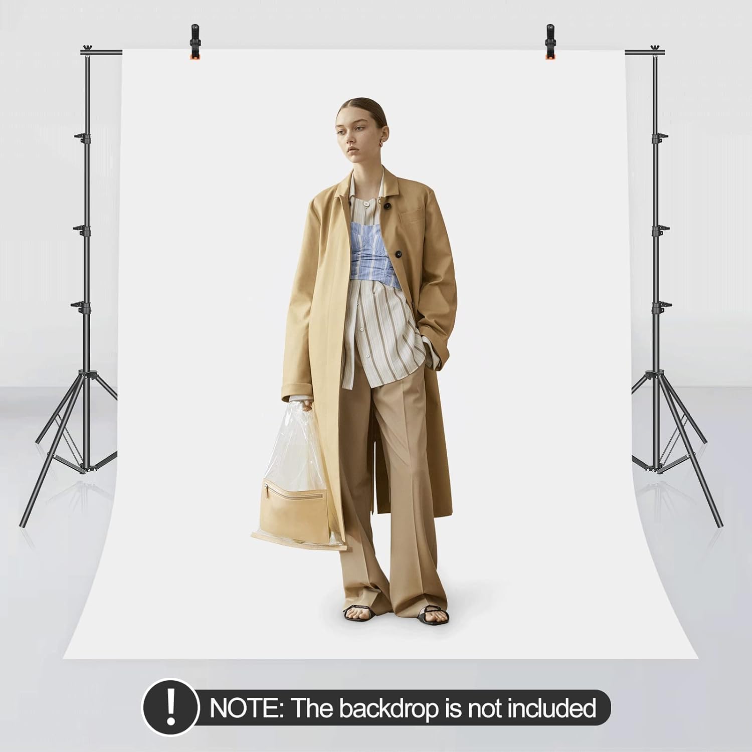 Model wearing a beige coat and pants with a white backdrop, holding a clear bag.