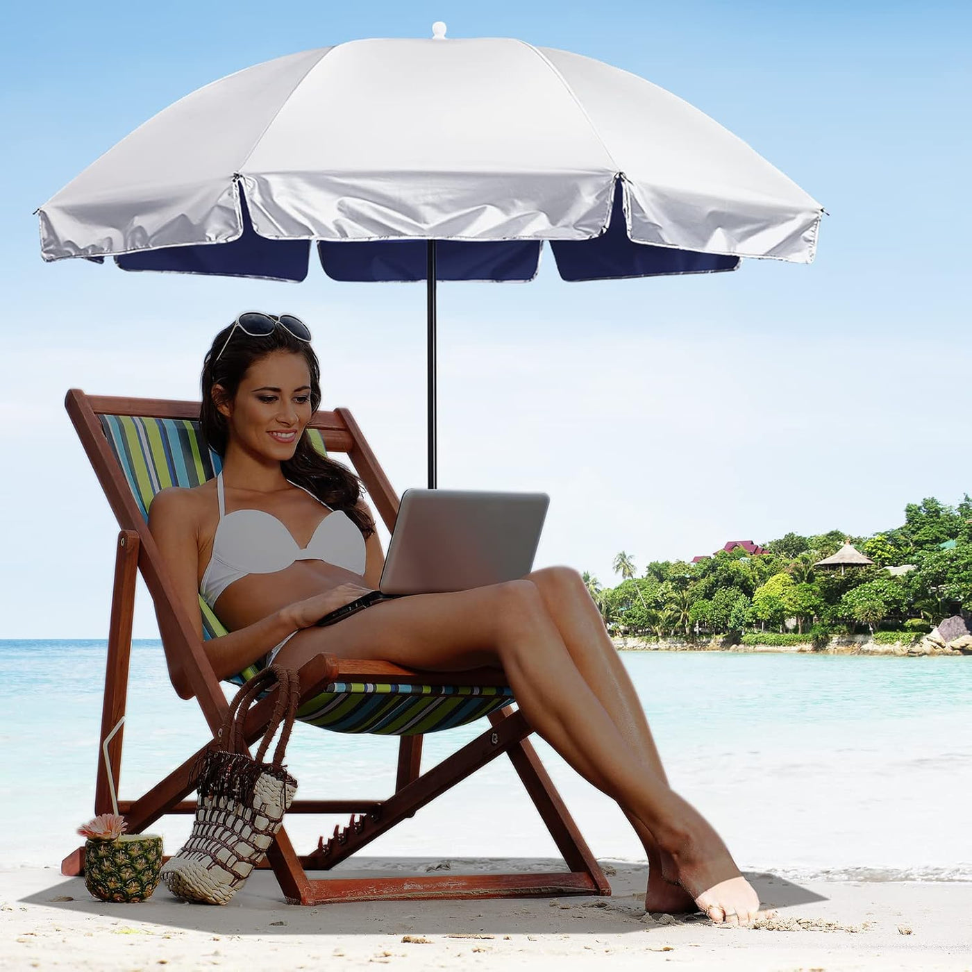 A woman relaxes in a striped beach lounge chair under a large white beach umbrella on a sandy beach, using a laptop. A woven bag and a small pineapple-shaped item sit beside the chair, with turquoise water and trees in the background.
