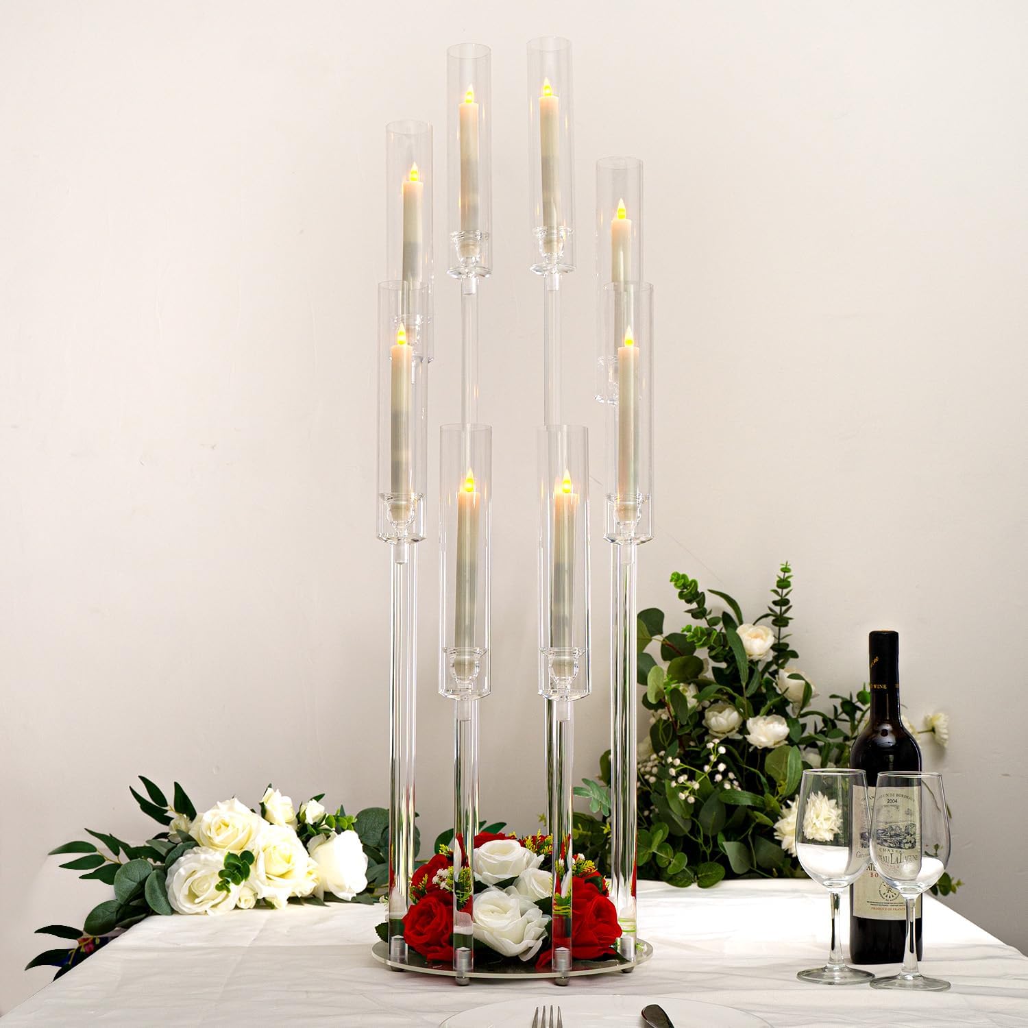 Decorative table setting with tall clear candles, flowers, and a bottle of wine on a white surface.