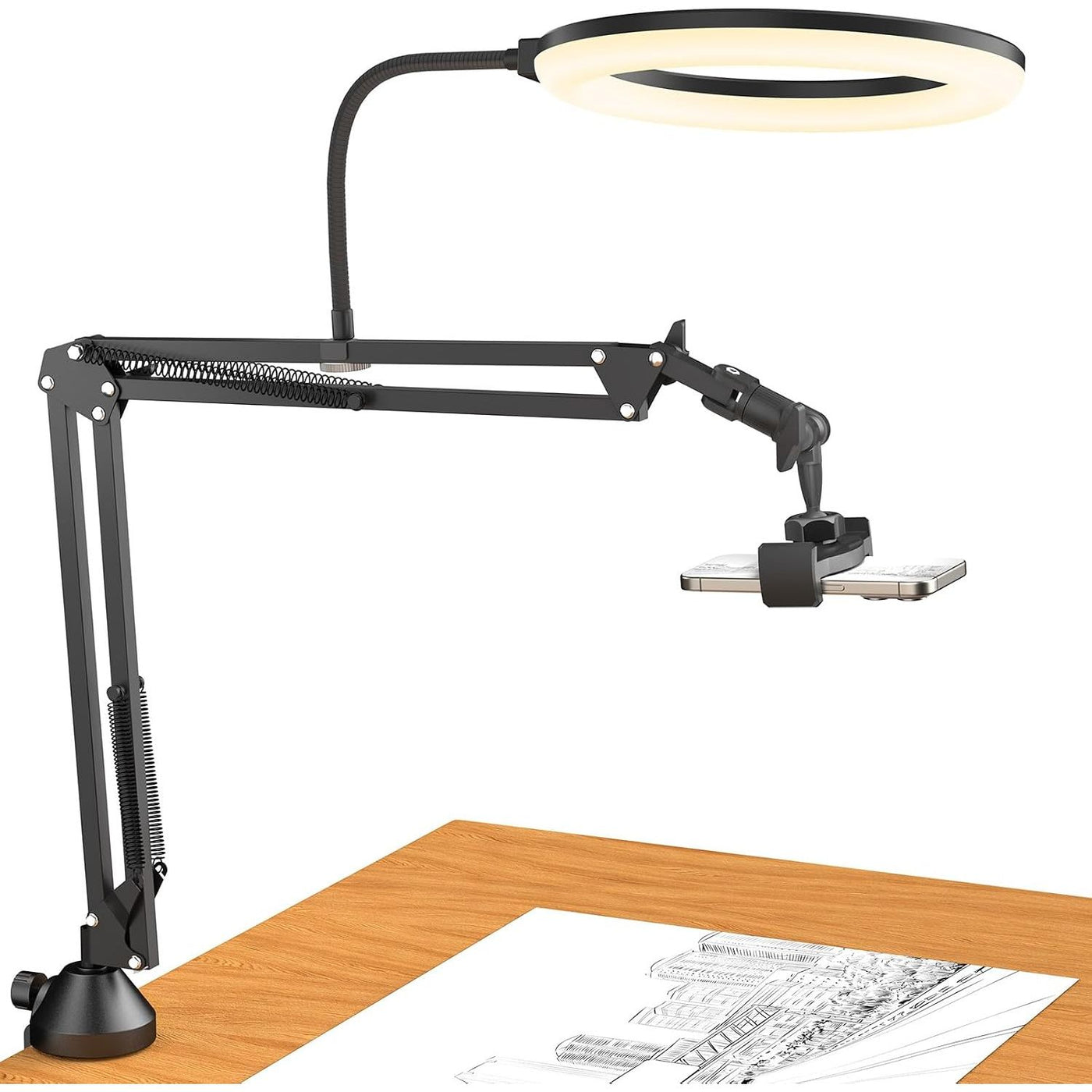 Black adjustable desk lamp with a circular LED ring light and articulated arm, shown mounted on a wooden desk with a phone clamp holding a smartphone and sketch papers below.