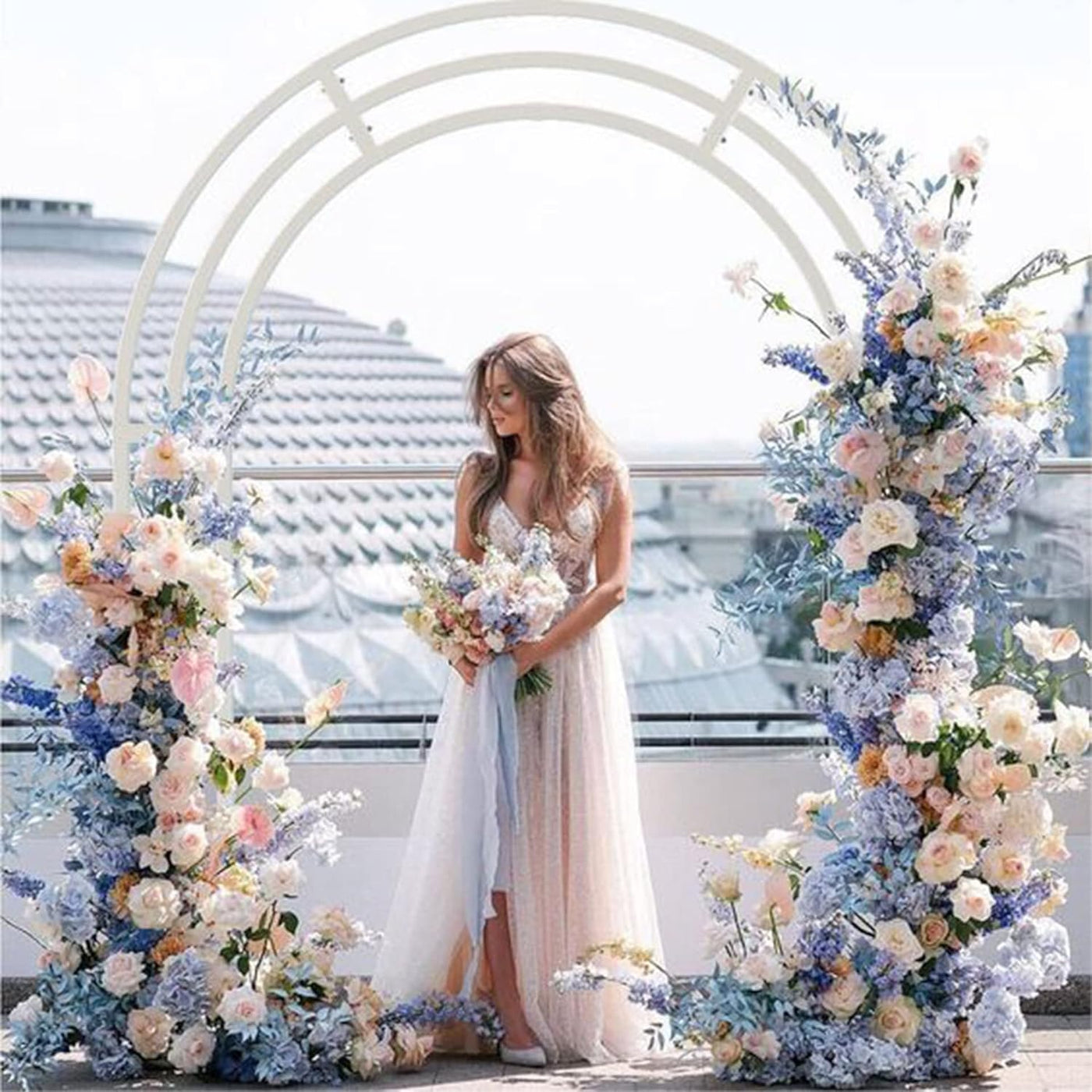 Woman in a wedding dress standing between two floral arches on a rooftop with a cityscape background.