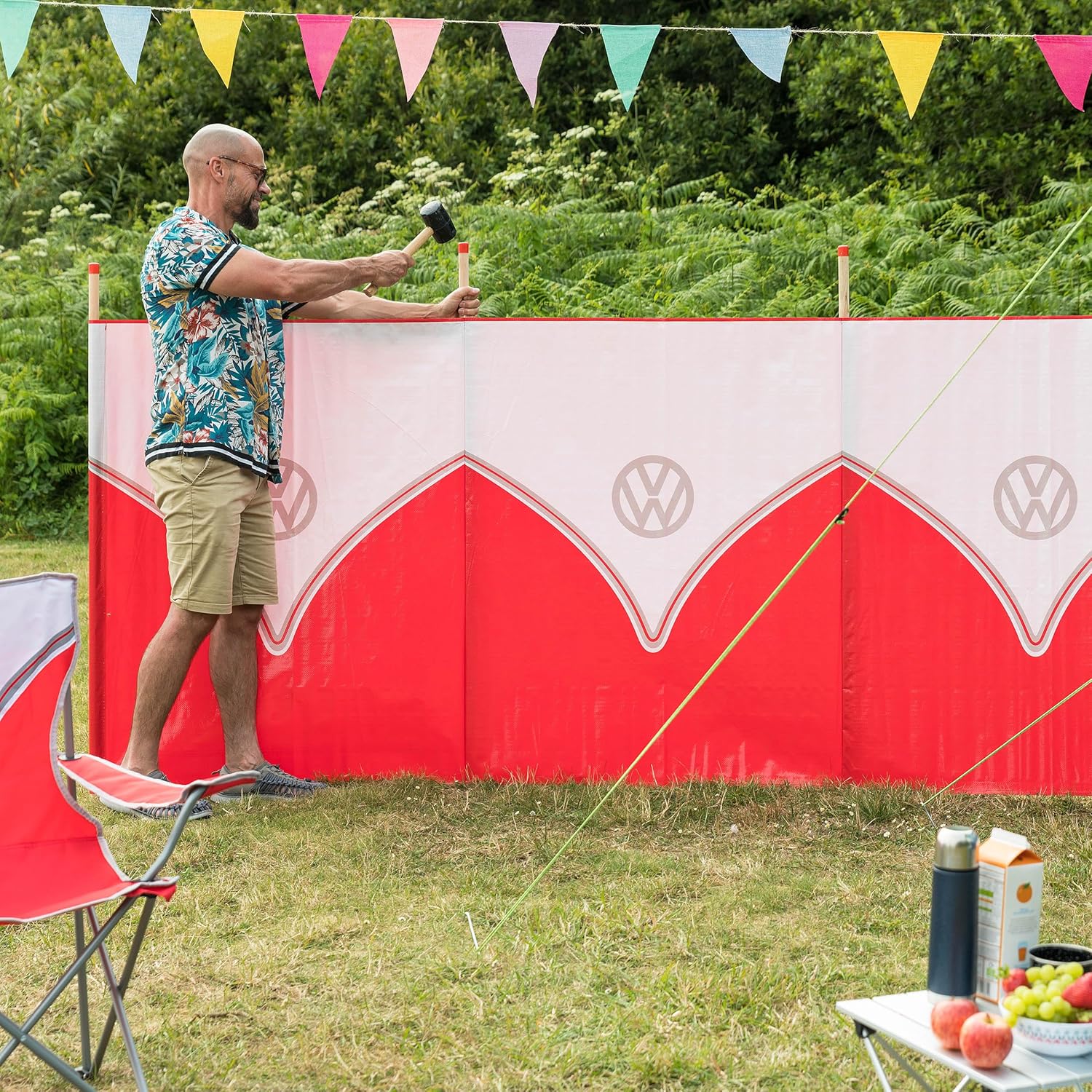 A man in a tropical print shirt and khaki shorts stands outdoors holding a mallet against a red and white VW-themed backdrop, with grass, trees, bunting, chairs, and a small table visible.