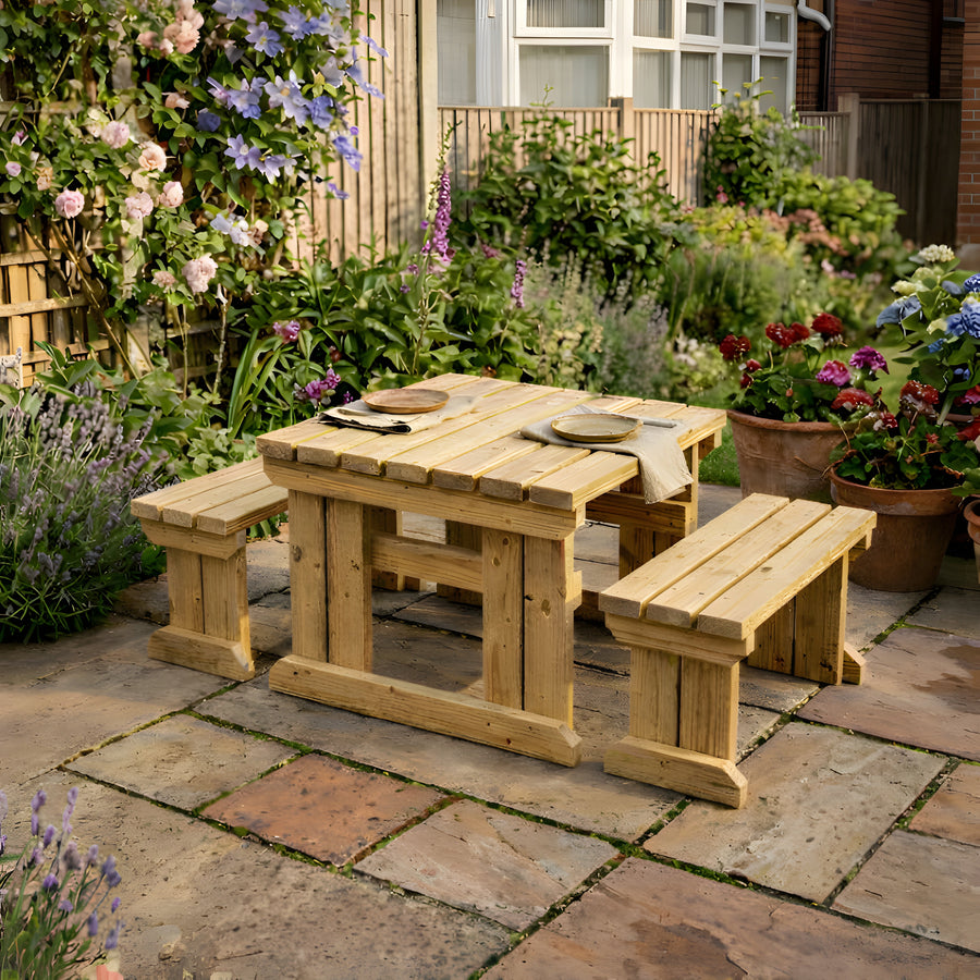 Wooden outdoor table with benches on a stone patio surrounded by garden plants.