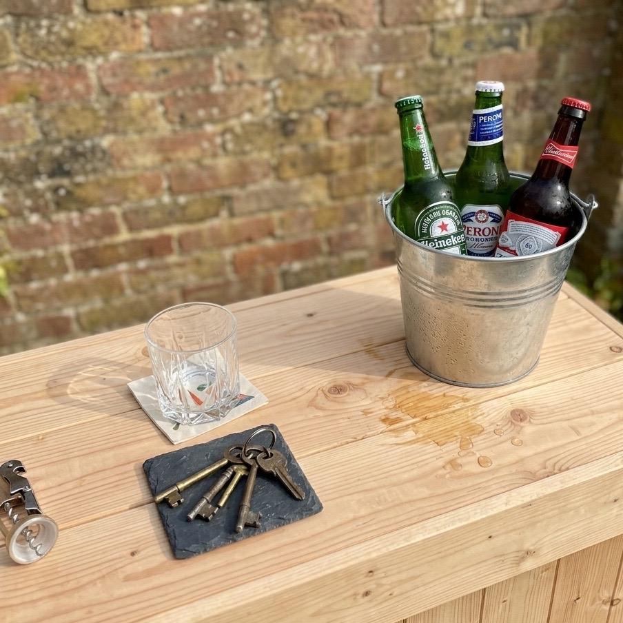 Three beer bottles in a metal bucket on a wooden table with a glass and keys.