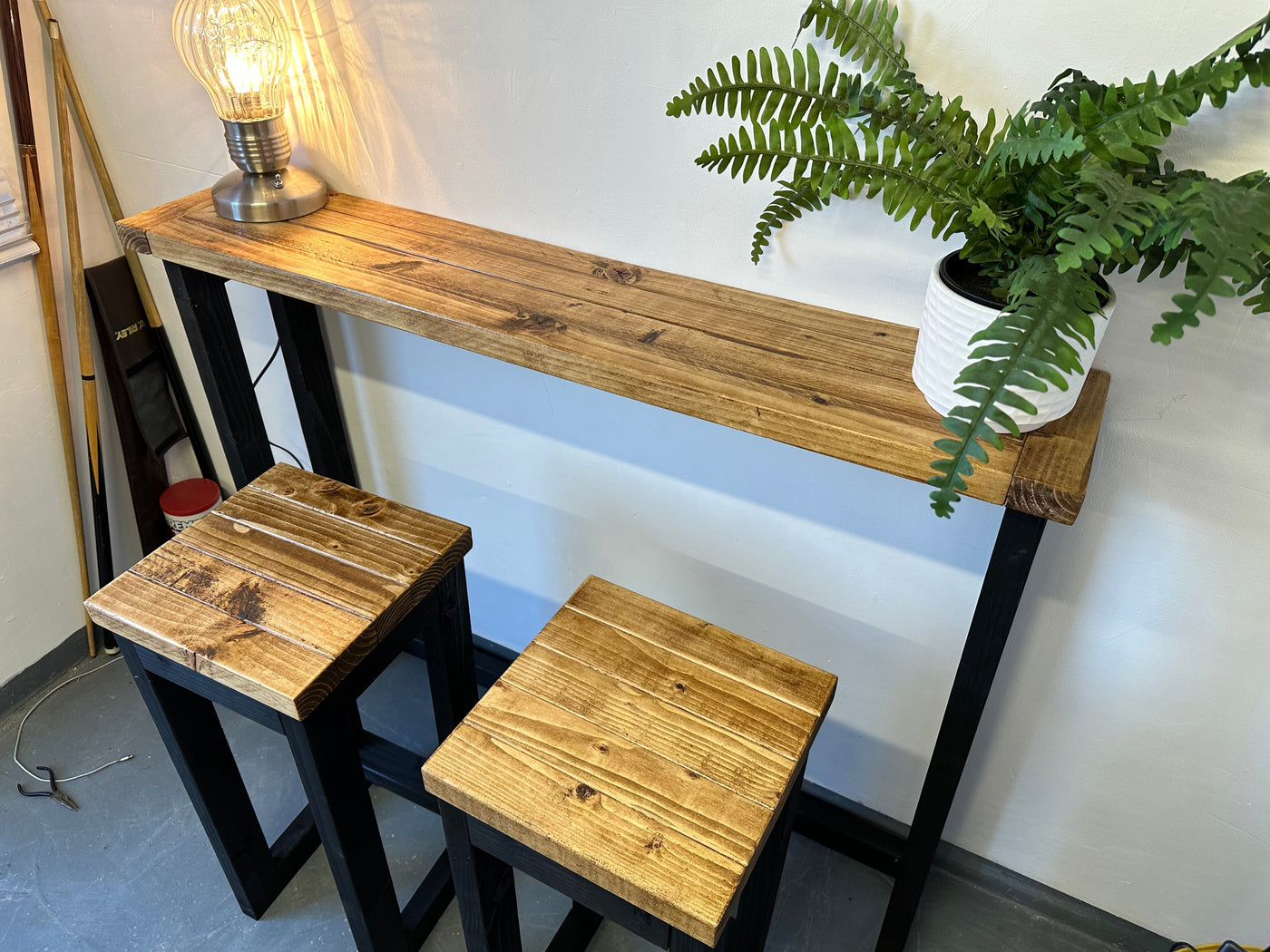 Wooden bar height table with two stools and a plant in the background.