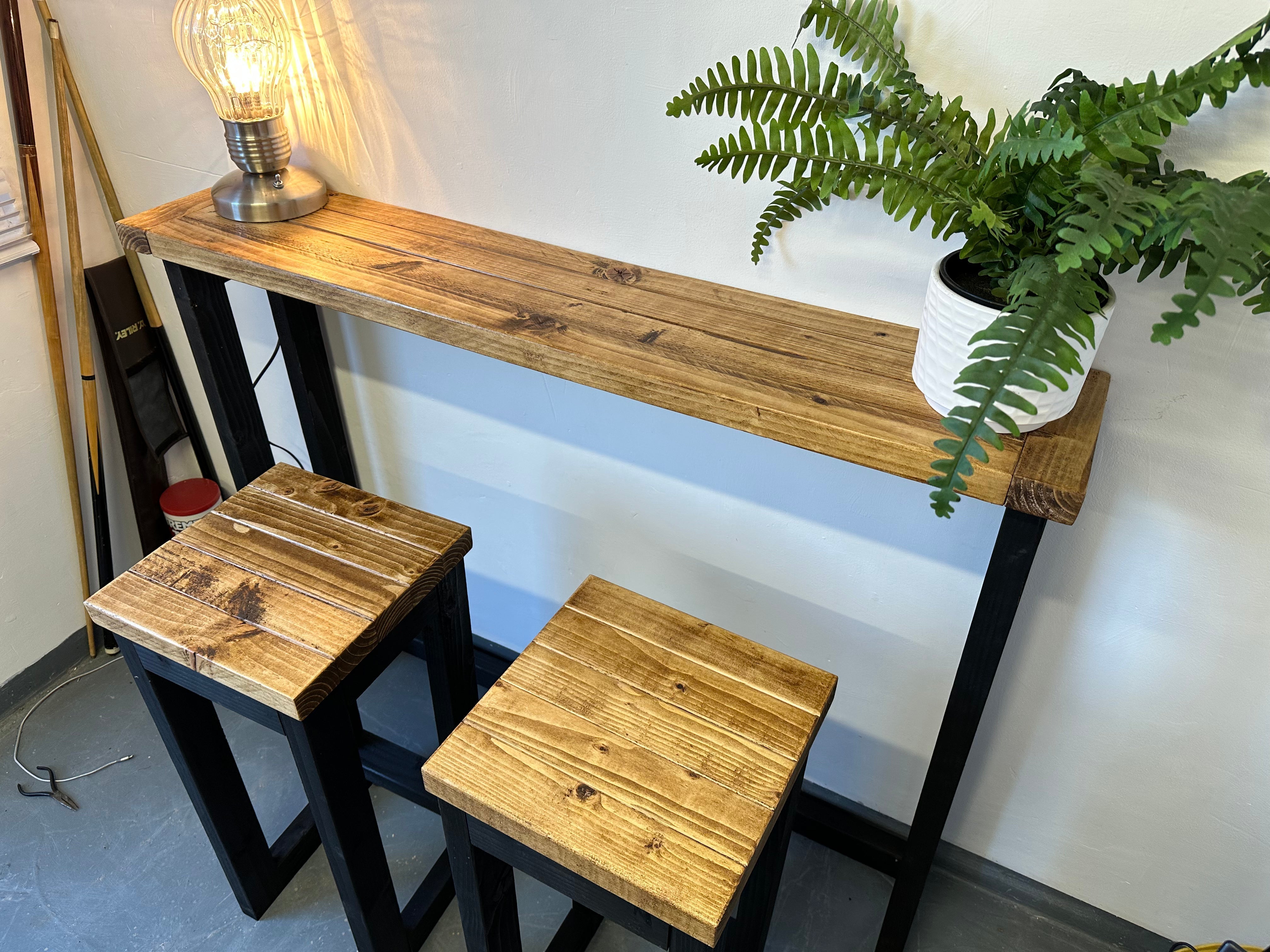 Wooden bar height table with two stools and a plant in the background.