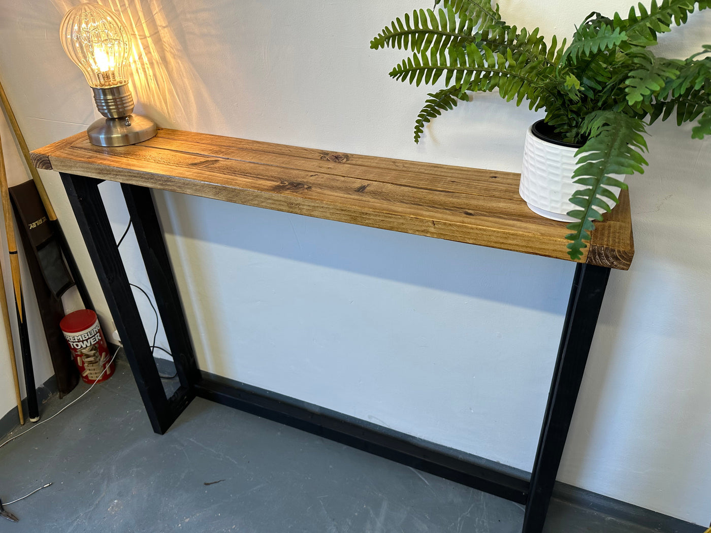 Wooden console table with black metal legs, lamp, and plant against a white wall.