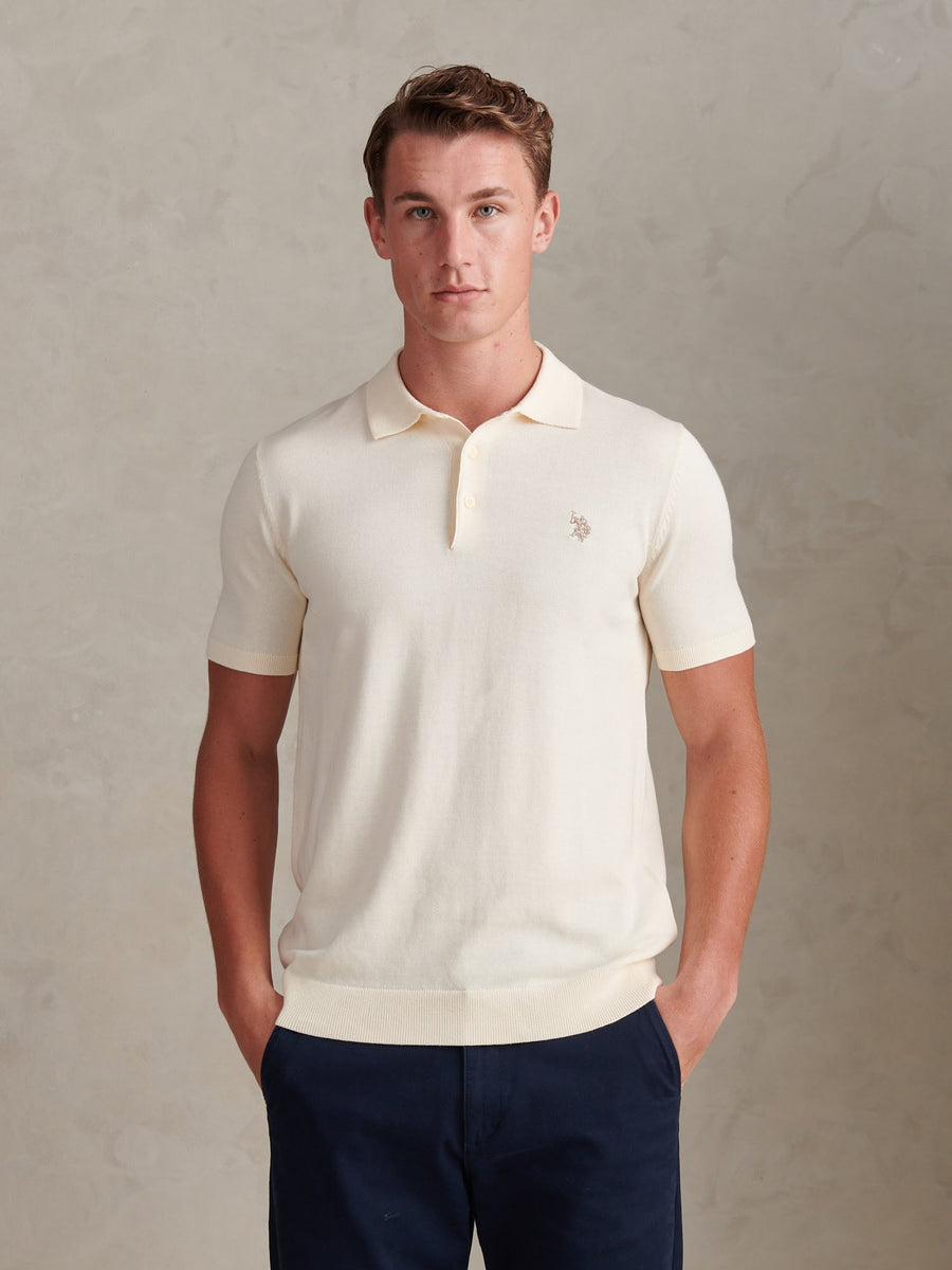 Man wearing a cream polo shirt with a logo against a plain background