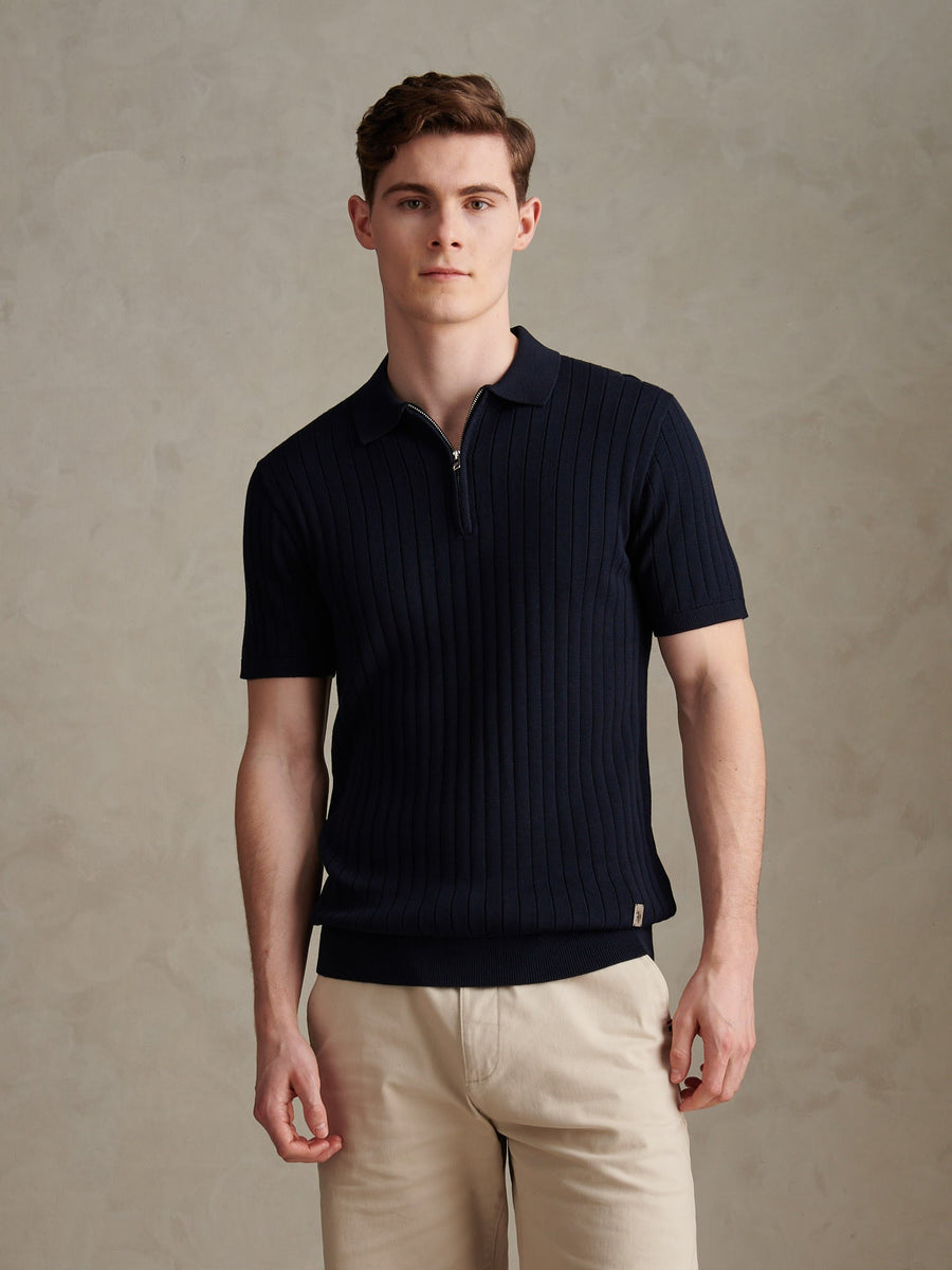 Man wearing a navy ribbed polo shirt and beige pants against a beige background
