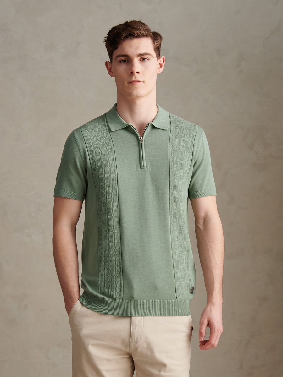 Man wearing a green polo shirt and beige pants against a plain background