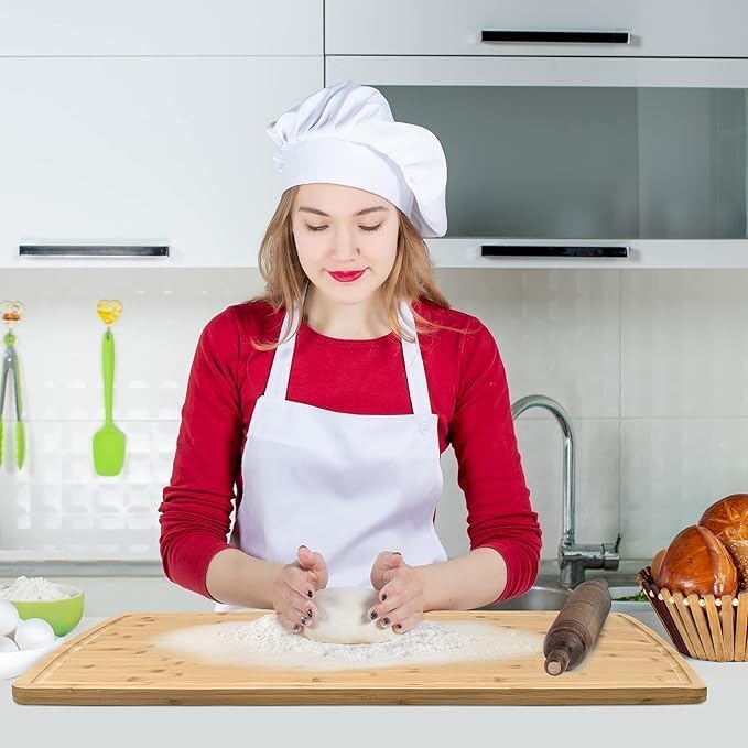 Woman in a kitchen wearing a chef's hat and apron, kneading dough on a wooden board.