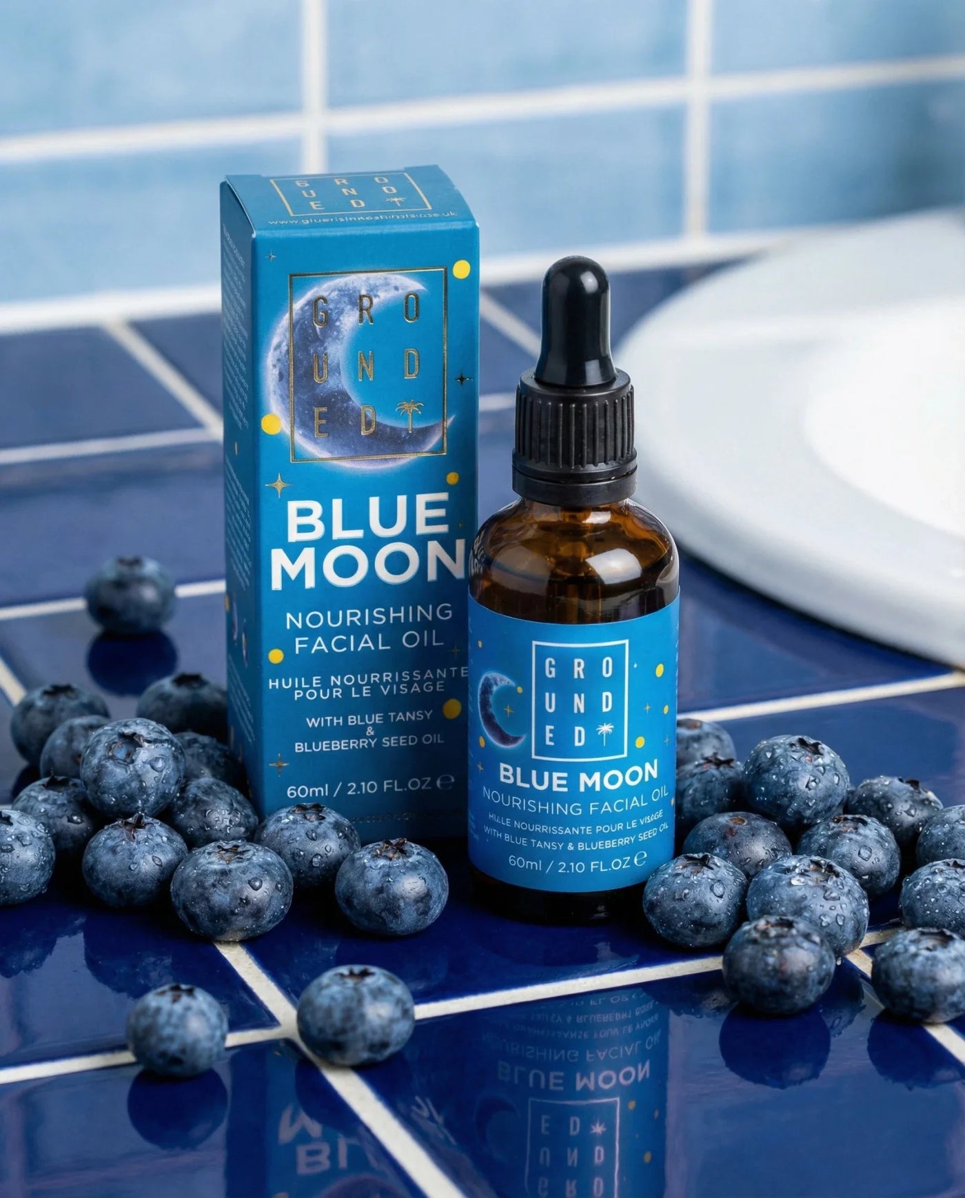 Blue Moon facial oil bottle and packaging on a blue tiled surface with blueberries.
