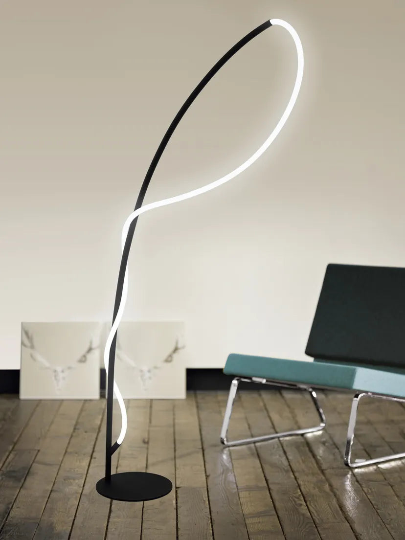 A modern black floor lamp with a glowing white curved LED light tube, standing on a dark wood floor in a minimalist room next to a teal chair.