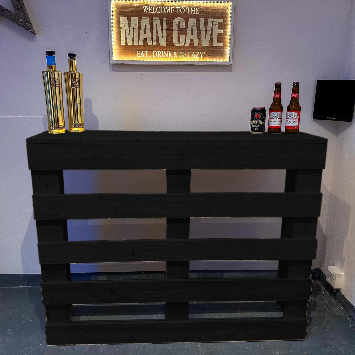 Man cave setup with a wooden pallet bar, bottles, and a neon sign.