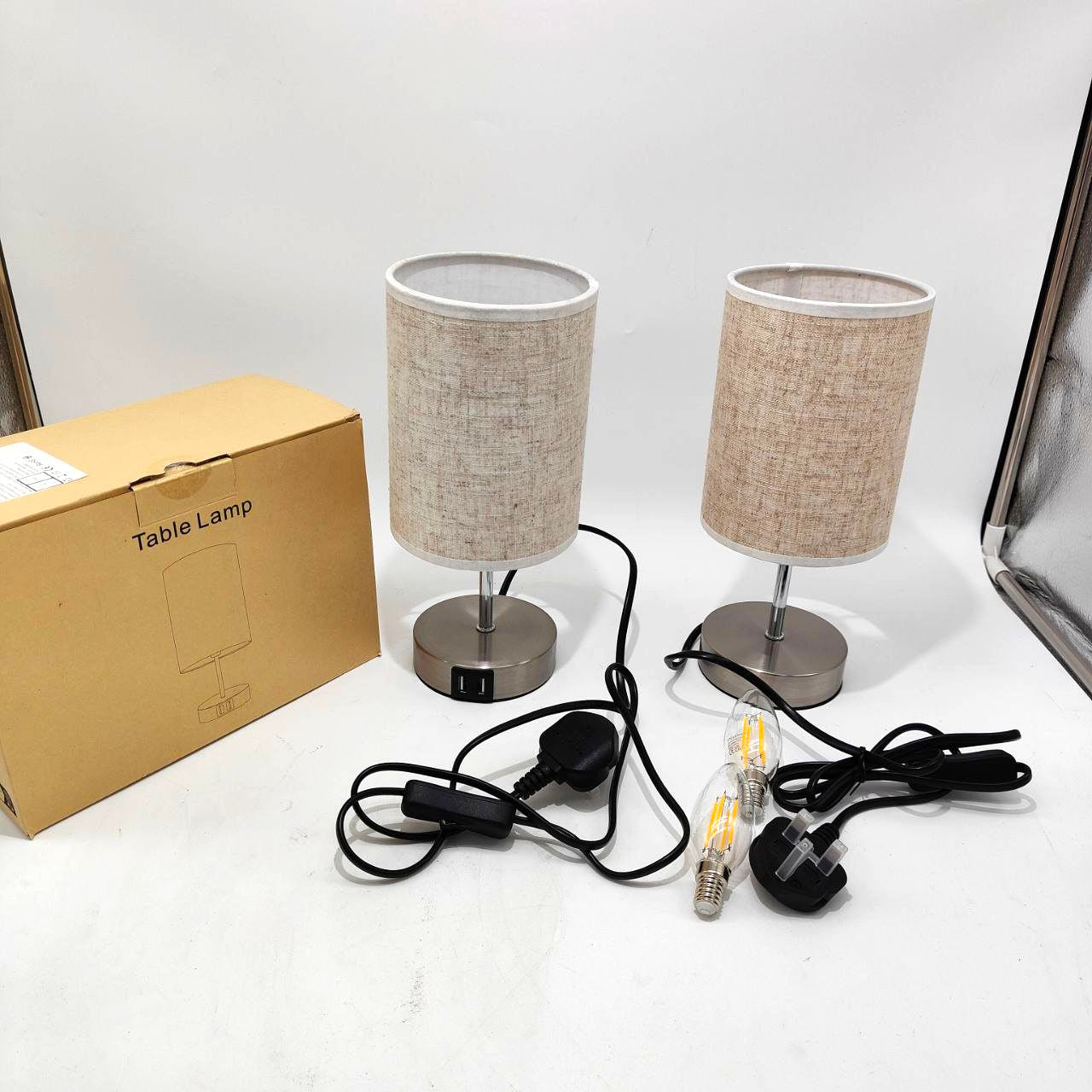 Two table lamps with beige fabric shades and metal bases, next to their packaging box on a white surface.