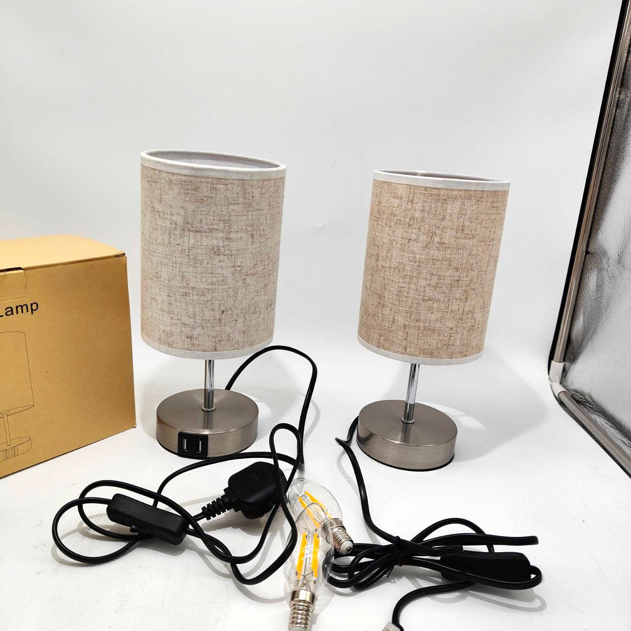 Two small table lamps with beige lampshades on a white surface, with a cardboard box labeled 'Lamp' in the background.