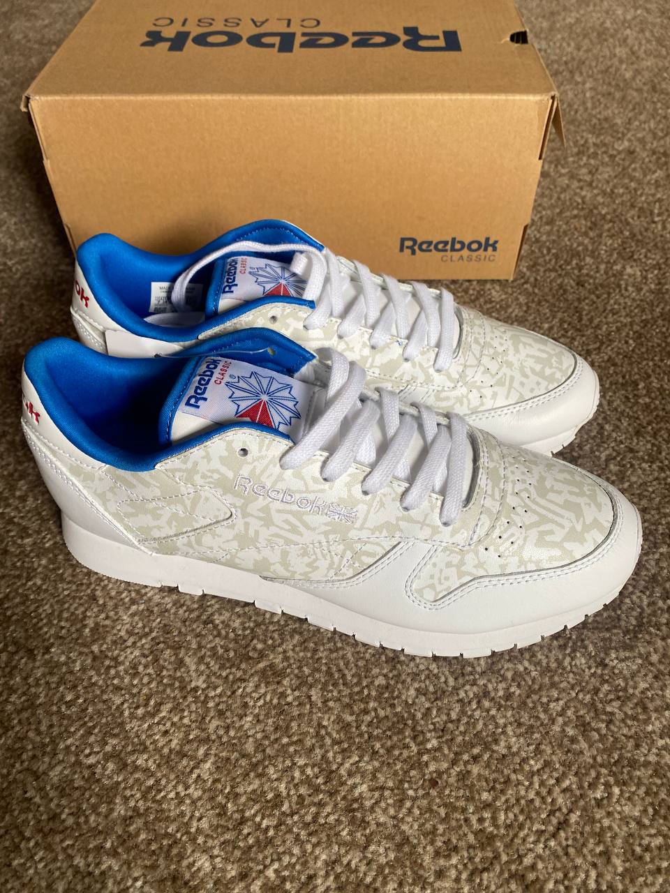White Reebok sneakers with blue accents on a brown carpet next to a Reebok box.