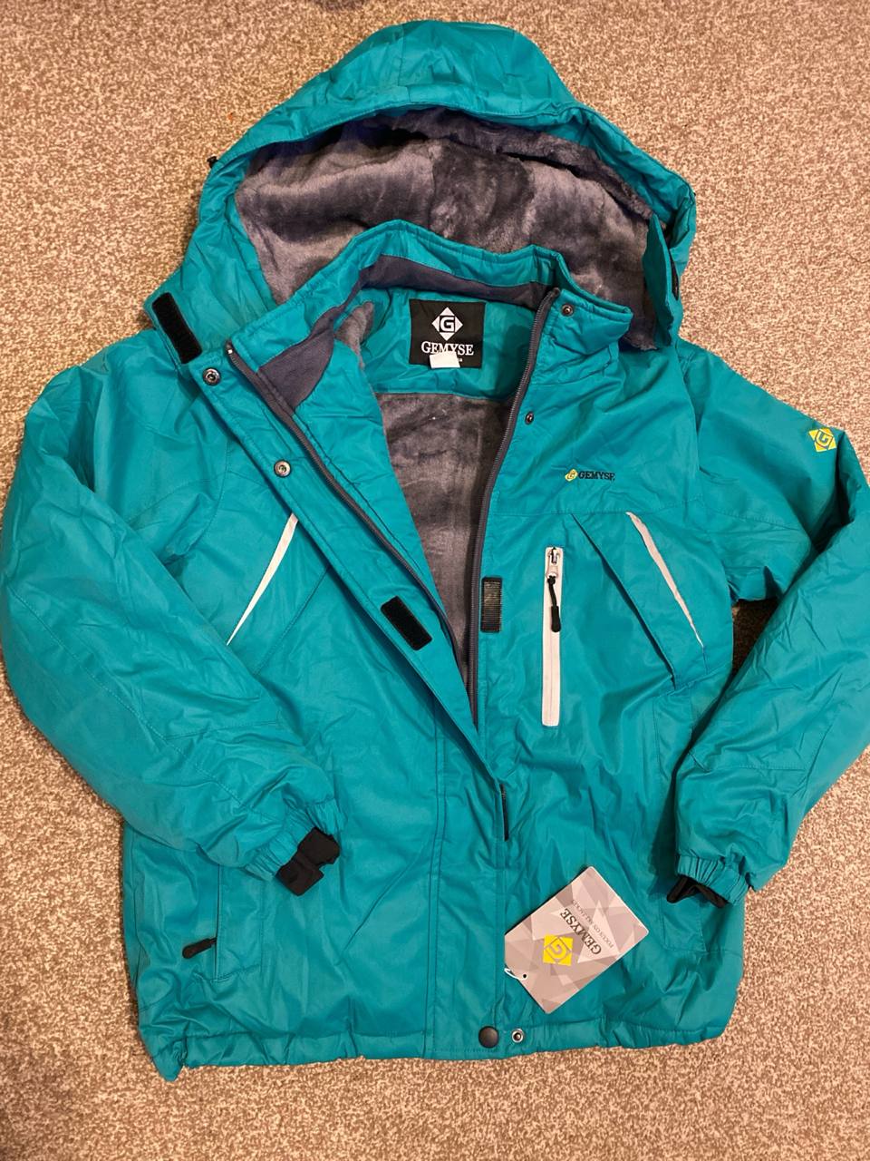 Teal jacket with brand logo on a beige carpet