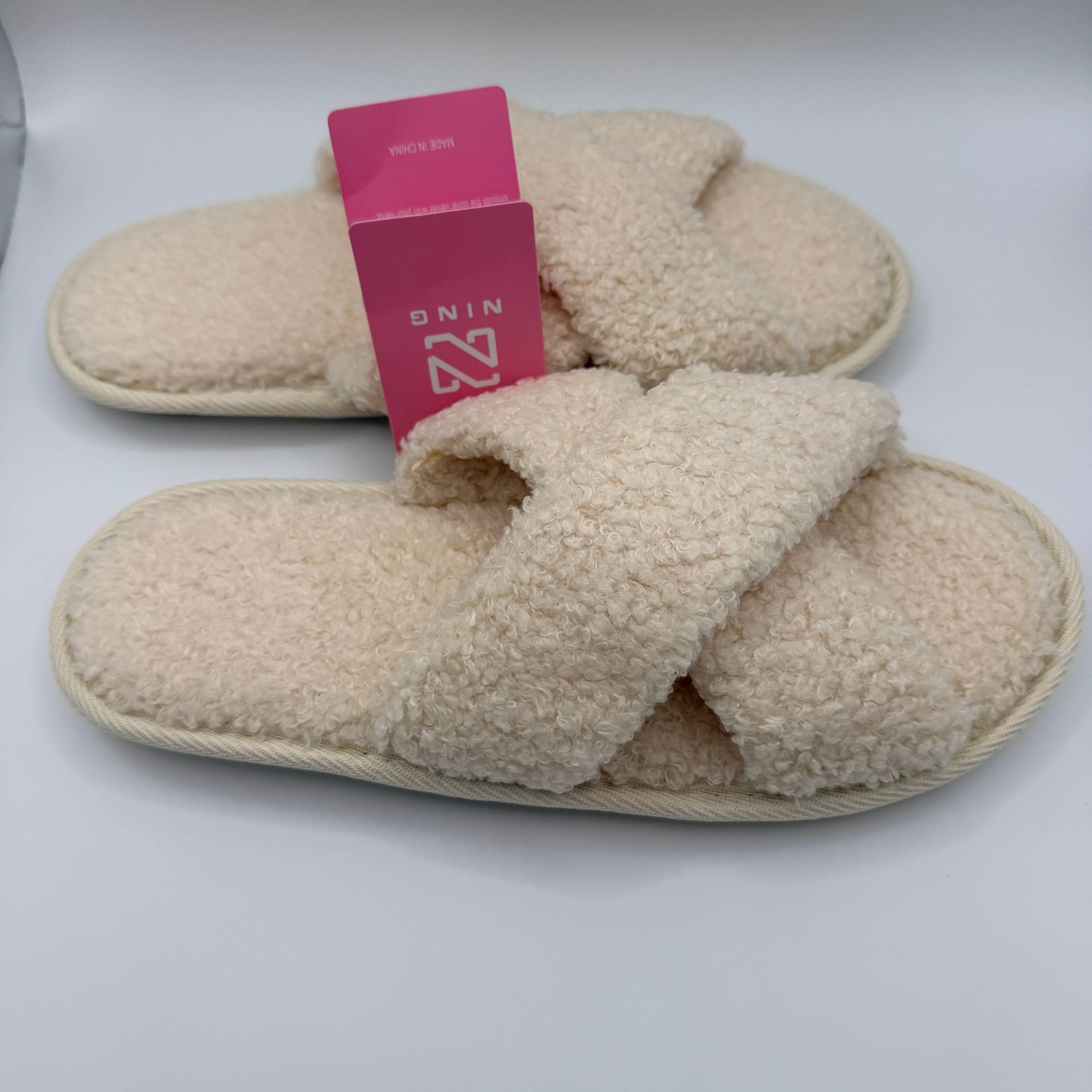 Pair of beige slippers with a pink tag on a white background