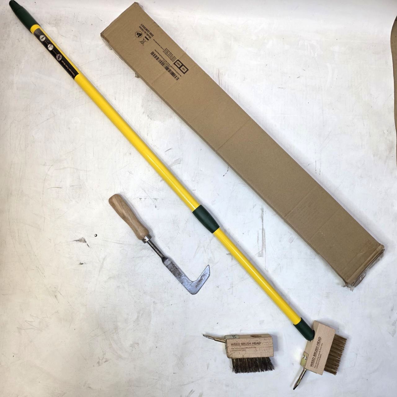 Cleaning tools including a long-handled brush and scraper with a cardboard box on a light surface.