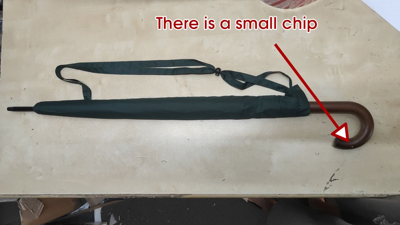 A black umbrella with a dark brown wooden handle lies on a beige tabletop. Red text at the top reads 'There is a small chip' with a red arrow pointing to a small damaged area on the wooden handle.