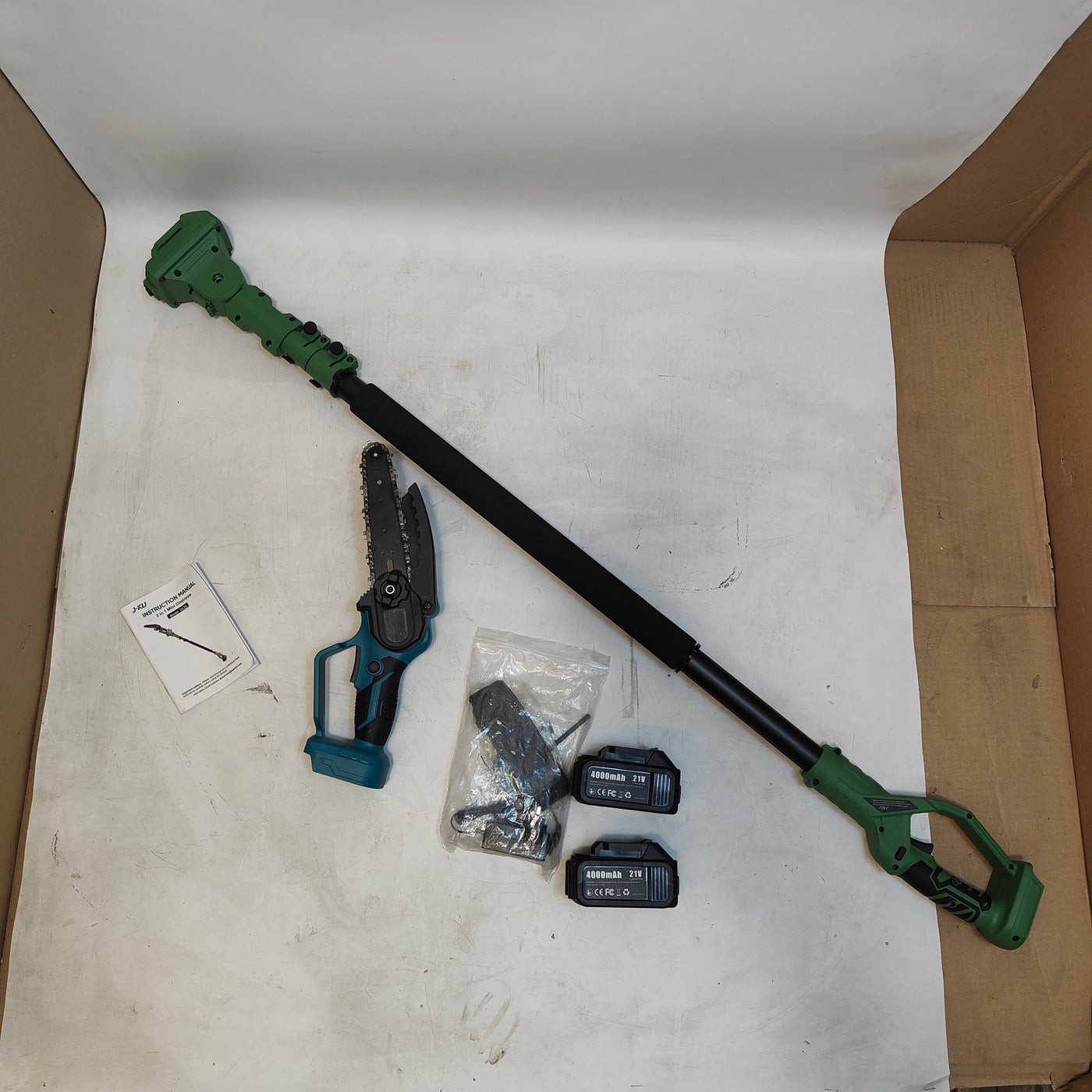 A cordless pole hedge trimmer with a telescopic black shaft, green trimmer head and handle, shown with a small hand saw, two black batteries, mounting hardware, and an instruction card inside an open cardboard box on a white studio sweep.