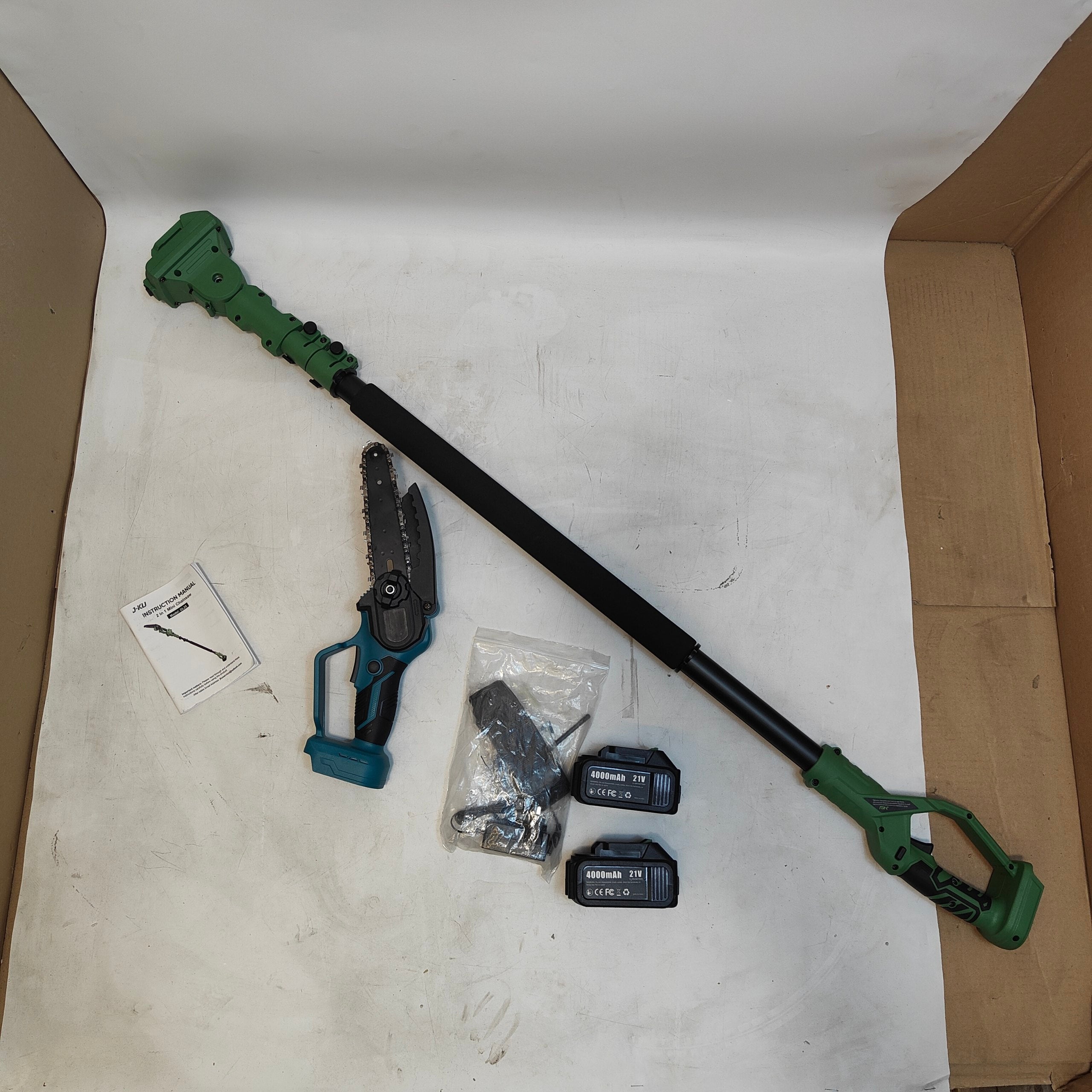A cordless pole hedge trimmer with a telescopic black shaft, green trimmer head and handle, shown with a small hand saw, two black batteries, mounting hardware, and an instruction card inside an open cardboard box on a white studio sweep.