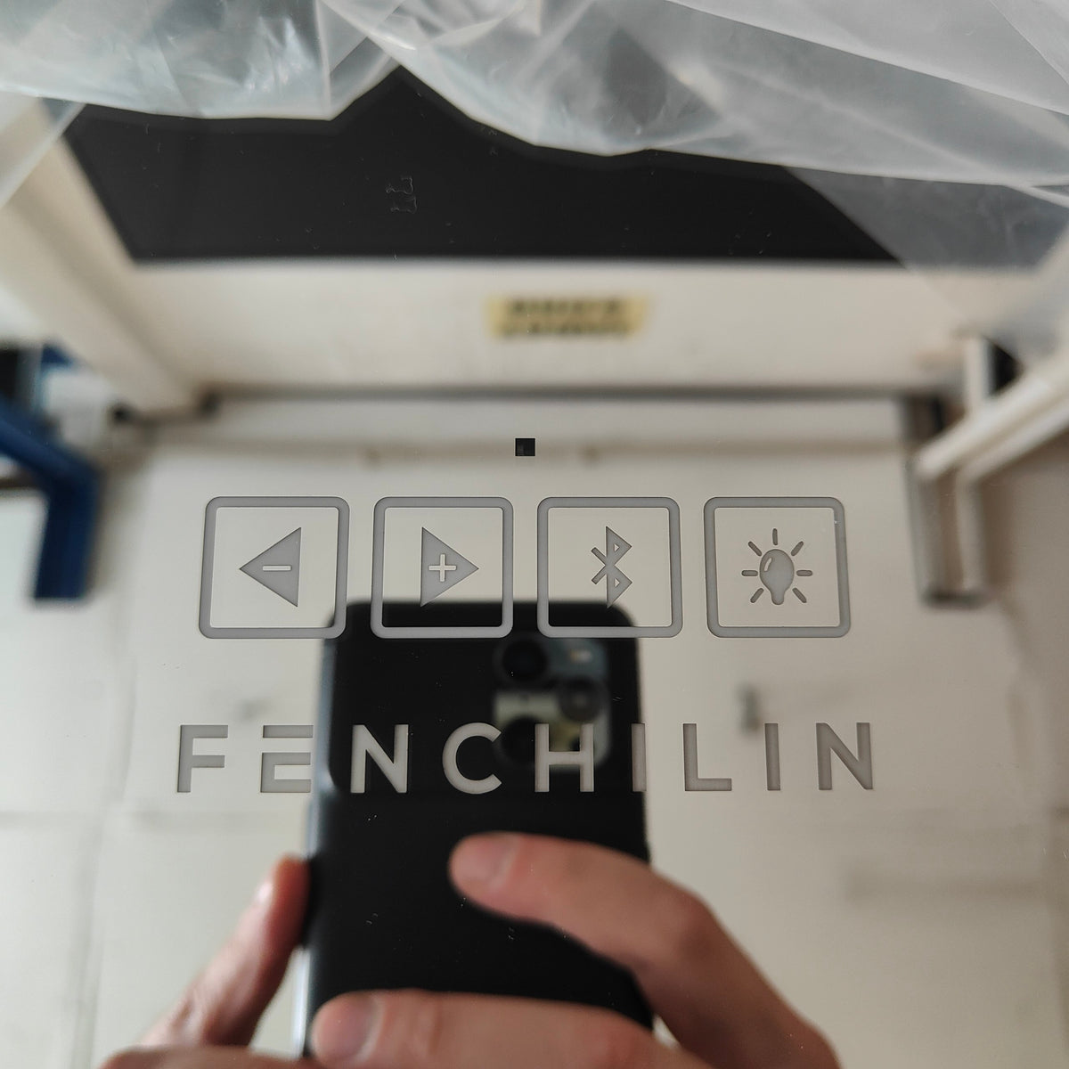 A close-up of a hand holding a black smartphone in front of a white surface, showing the back of the phone with a frosted or tinted film. The film has a translucent gray design with icons for back, play, Bluetooth, and brightness, plus the word FENCHILIN.