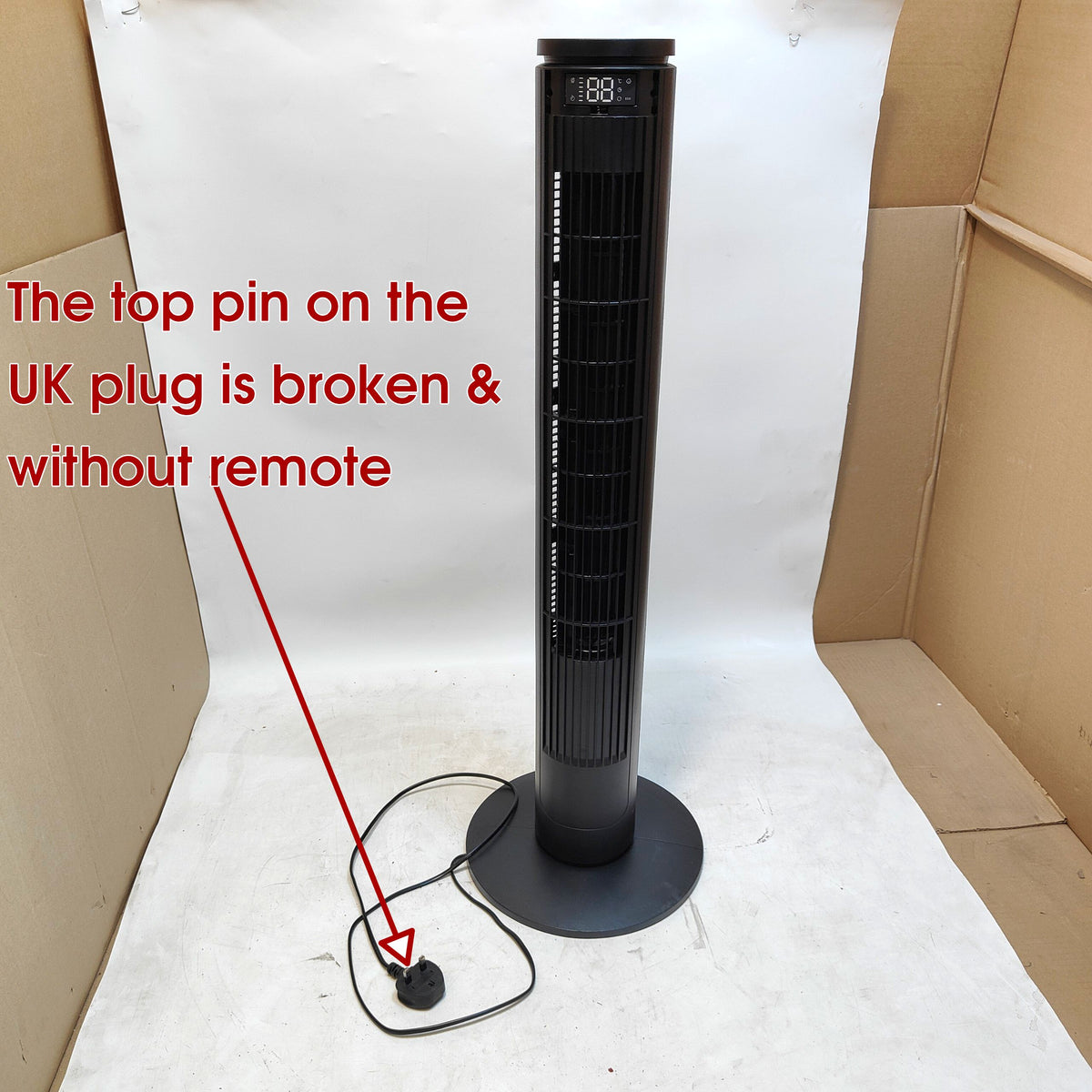 A tall black tower fan standing upright on a white backdrop inside a cardboard box. Red text points out that the top pin on the UK plug is broken and there is no remote.