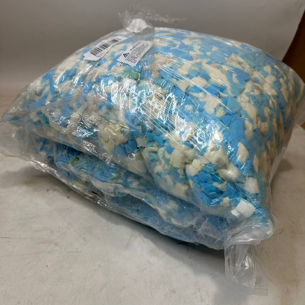 A large clear plastic bag filled with blue and white foam or crumb material, photographed on a light gray surface against a light gray background.