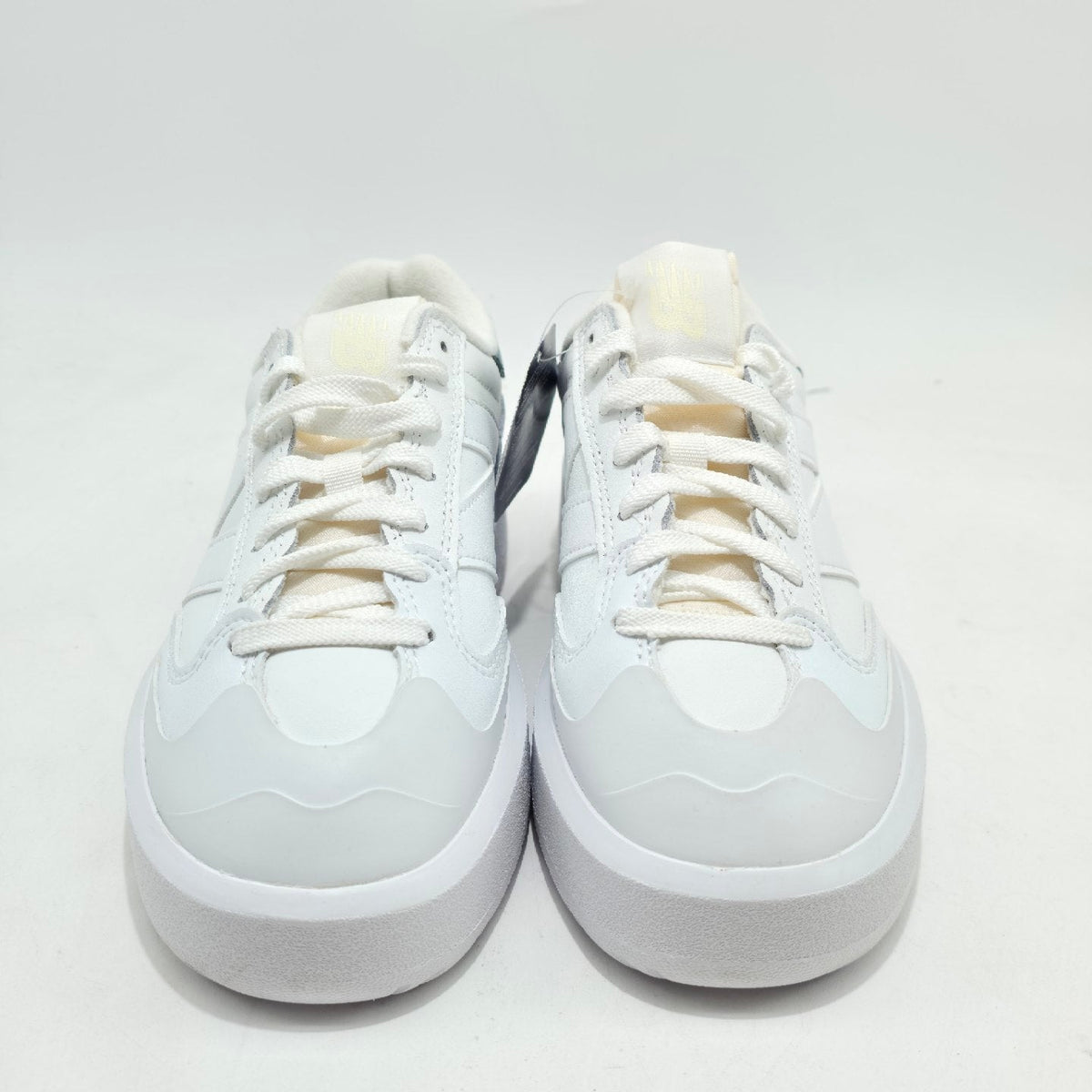 A pair of low-top white sneakers with white laces and thick white soles, photographed from the front on a plain white studio background.