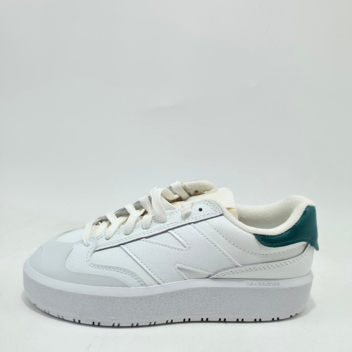 A white low-top sneaker with a chunky white sole, featuring a small dark green heel tab accent.