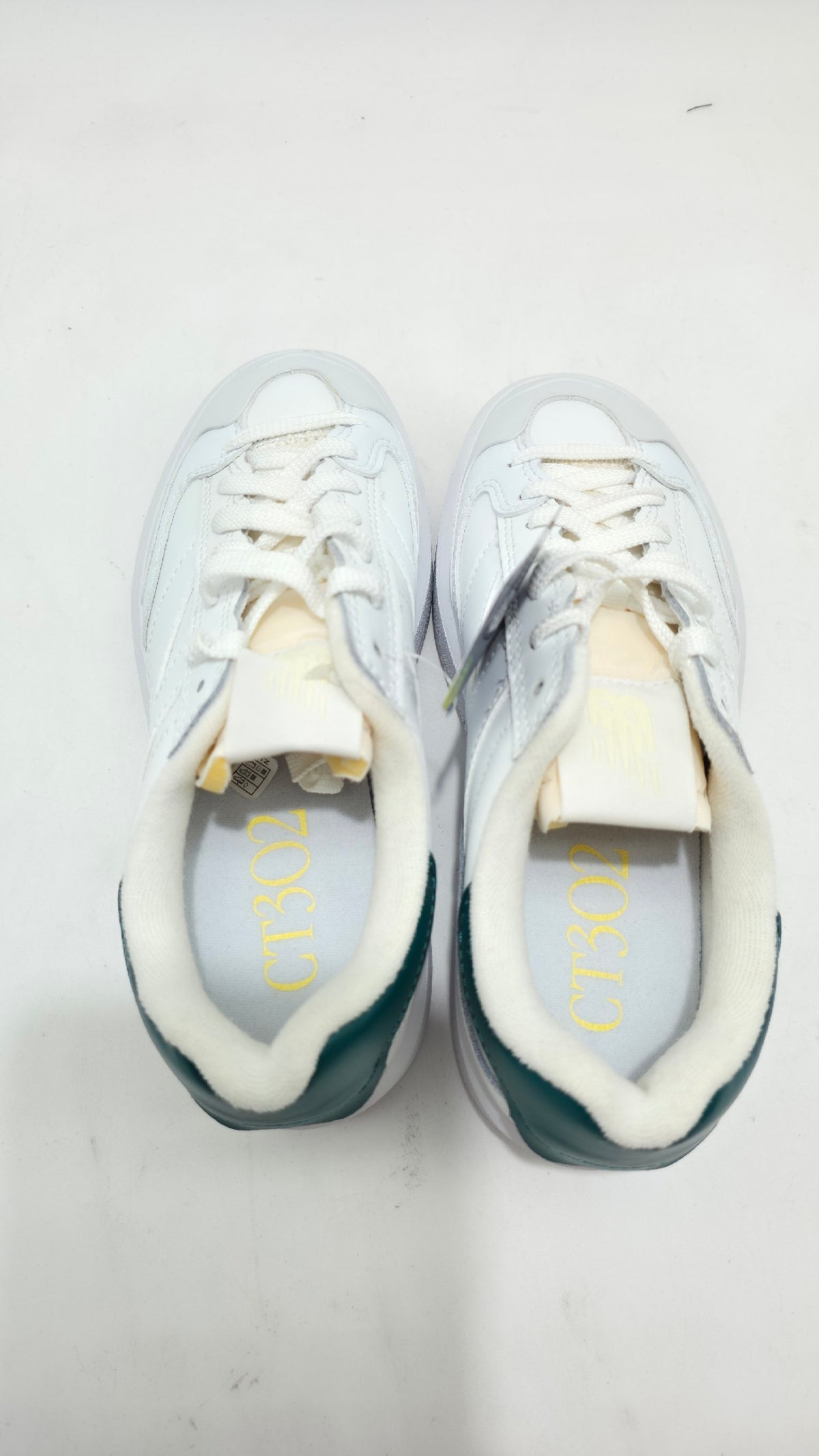 A pair of white low-top sneakers viewed from above, featuring white laces, a white faux-fur collar lining, and dark teal-green heel accents with gold lettering inside the soles.